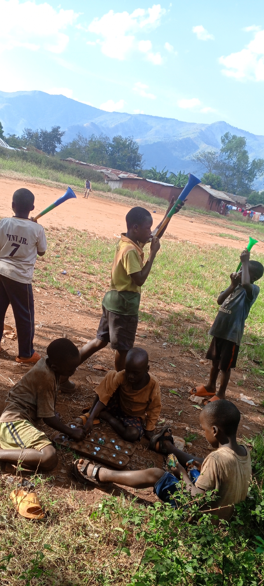 A group of young boys are gathered in an outdoor area in Ngalula, Democratic Republic of the Congo. Four are seated on the ground, playing a game with marbles on a wooden board. Three other boys are standing, each holding a blue and green plastic horn or vuvuzela. Mountains are visible in the background. The ground is dirt, with patches of grass and weeds. Several simple buildings are present in the background.