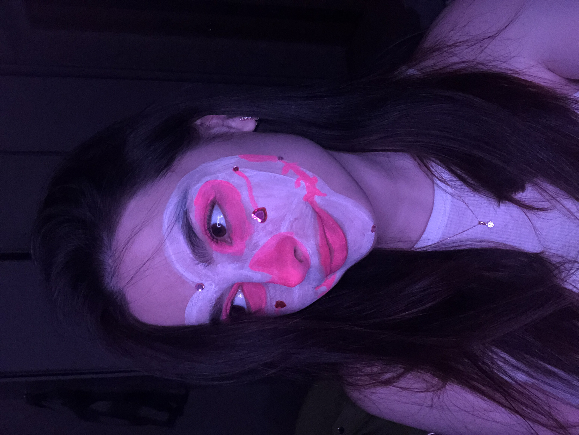 The image displays a close-up portrait of a person, likely a young woman, in a low-light indoor environment, bathed in purple illumination characteristic of a blacklight or UV setting. Her face is adorned with intricate face paint, featuring a white base with bold, vibrant hot pink accents forming abstract shapes and highlighting facial features. Small, shimmering rhinestones or gems are placed on her forehead, above her eyes, and on her chin. She has long, dark hair cascading around her face and shoulders. The subject is wearing a white ribbed top or tank top, and a delicate necklace with a small, sparkly pendant (possibly star-shaped or floral with a central gem) is visible. Her eyes are open and gaze slightly away from the camera. This artistic makeup suggests a themed event, party, or performance taking place at night in Noviy, Russia. There is no visible text in the image.