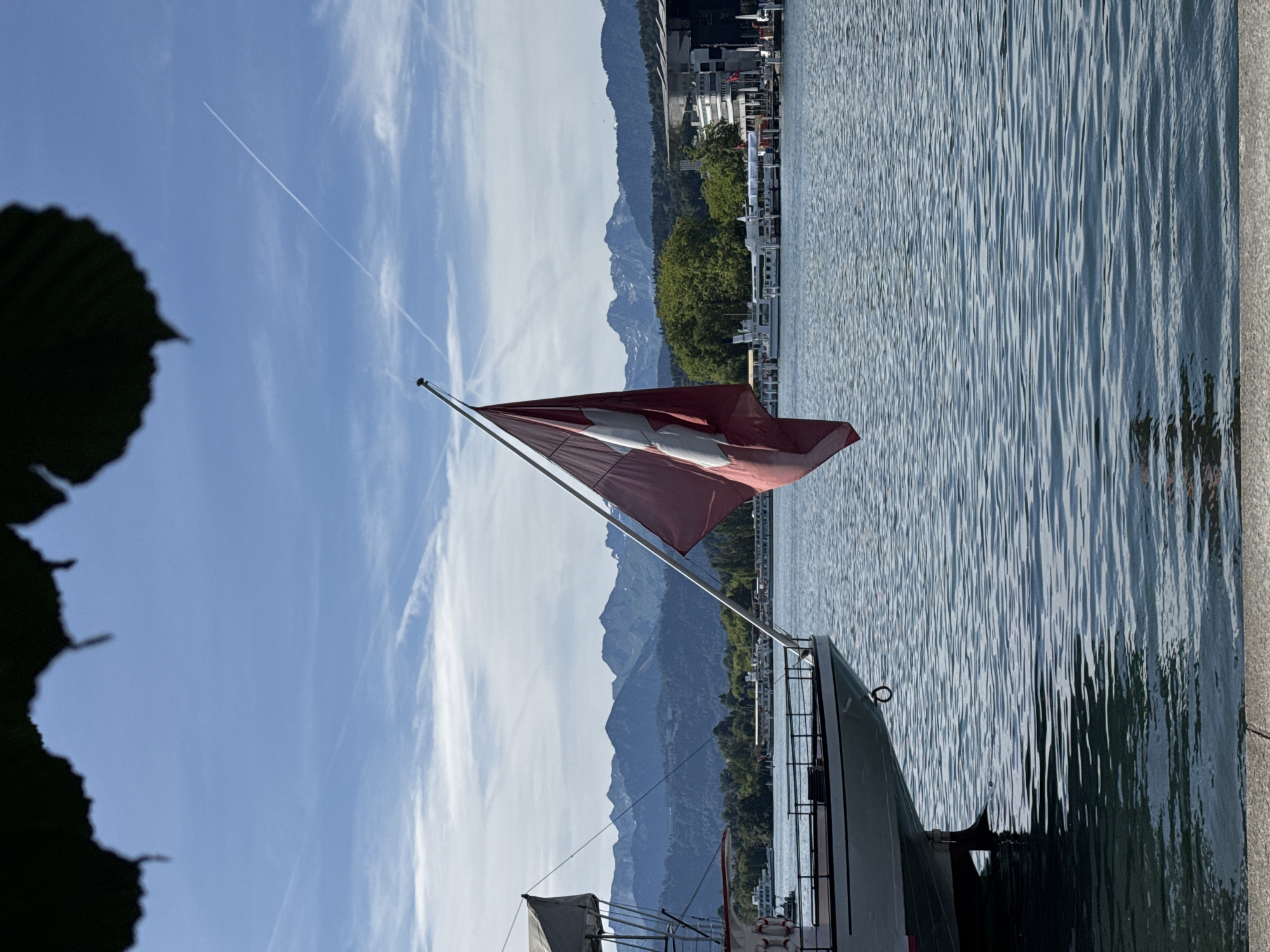 An outdoor, vertical shot captures a Swiss flag fluttering on a boat in the foreground, with a calm lake and mountains in the background. The flag is a deep red with a white cross in the center and is attached to a metal pole. The boat, white with red trim, is partially visible on the left side of the image, with a railing and a covered area. The lake's surface is rippled, reflecting the blue sky. In the distance, a range of blue-toned mountains forms the horizon, partially obscured by green trees. Buildings with modern architecture are visible along the lakeshore. The sky is a clear blue with thin, white contrails from airplanes. The upper part of the image is framed by dark, out-of-focus foliage. The scene is likely the city of Lucerne, Switzerland, given the iconic view of the lake and mountains.