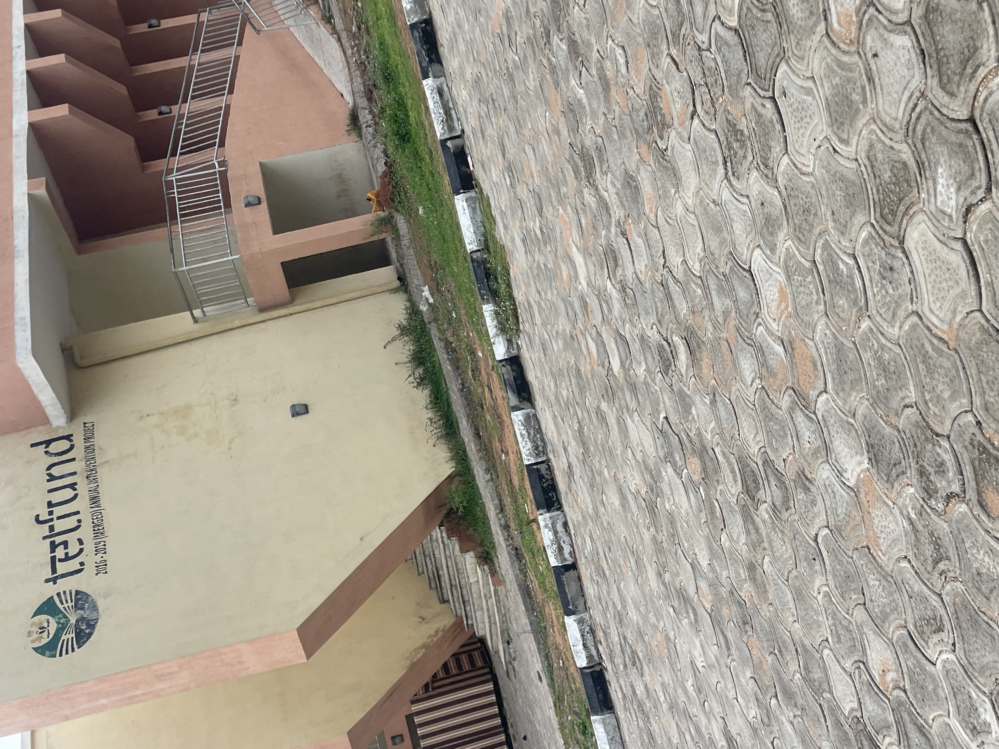 The image shows a building with a sign that reads "TETFUND 2016-2019 (MERGED) ANNUAL INTERVENTION PROJECT". The building is painted in shades of yellow and pink. There is a paved area in front of the building, with interlocking concrete blocks. The scene is located in Lokoja, Nigeria. There are no people visible in the image. The environment appears to be an outdoor setting, possibly a government or educational institution, given the signage. The architecture suggests a modern building design.