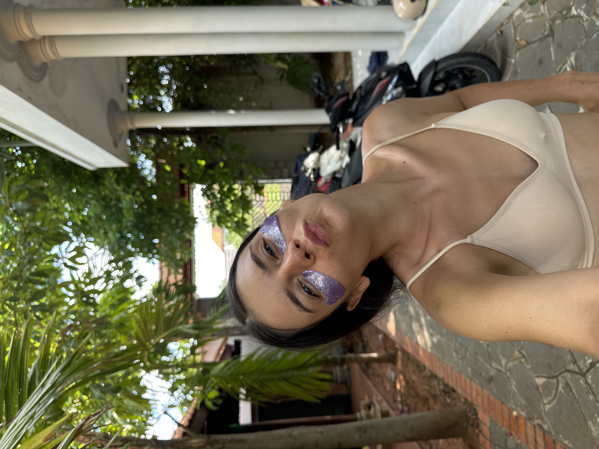 A young woman with dark hair and light skin is taking a selfie outdoors in Mũi Né, Vietnam. She is wearing a beige camisole and has purple glittery eye patches under her eyes. She is puckering her lips and tilting her head slightly. Behind her, a paved walkway leads to a building with white columns. A motorcycle is parked to the right of the woman. Lush green foliage and trees are visible in the background, suggesting a tropical or subtropical environment.