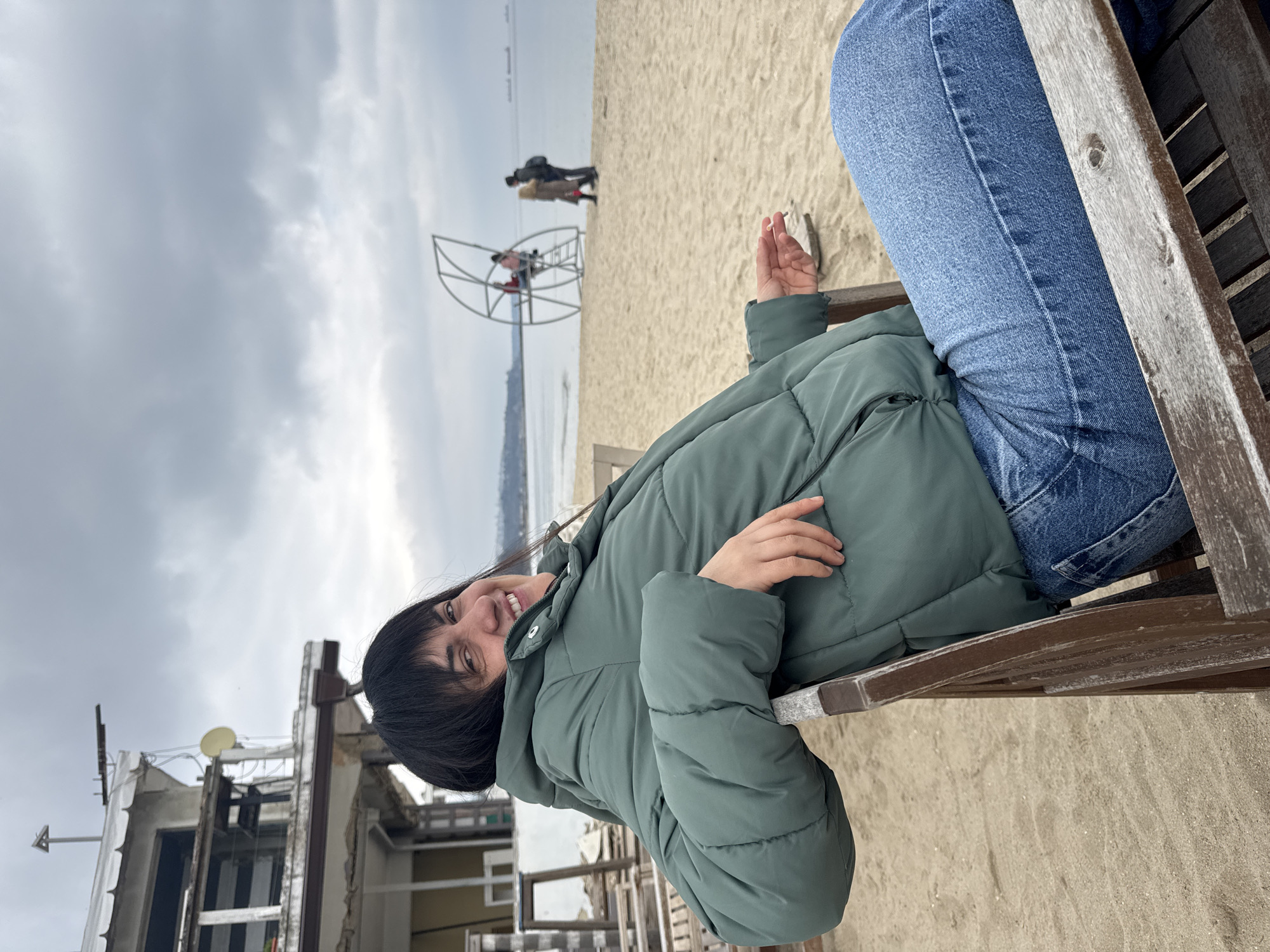 A medium shot shows a person reclining in a wooden beach chair on a beach in Varna, Bulgaria. The person is wearing blue jeans and a green puffer jacket and is smiling at the camera. The overcast sky illuminates the beach. A steel sculpture is visible on the beach, with a few pedestrians walking in the background. Faded beach structures are located to the left of the beach chair. The ground consists of light-colored sand.