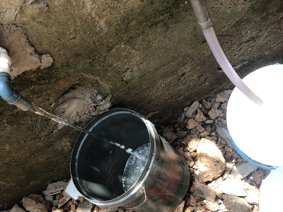 In the city of Alor, Nigeria, an outdoor scene depicts water collection from two distinct sources. In the upper left quadrant, clear water flows from a grey pipe elbow, connected to a white pipe, into a dark-colored metal bucket positioned below. This bucket, marked with red characters on its side, is partially filled with water. To the lower right, a translucent hose, originating from an unseen source, extends into a white bucket with a blue rim, also indicating water collection. The immediate ground surface is composed of scattered concrete debris and rocks. A textured, weathered concrete wall, exhibiting patches of darker discoloration, forms the background on the right side of the image. The scene is illuminated by natural light, casting subtle shadows among the debris. No individuals are present in the frame.