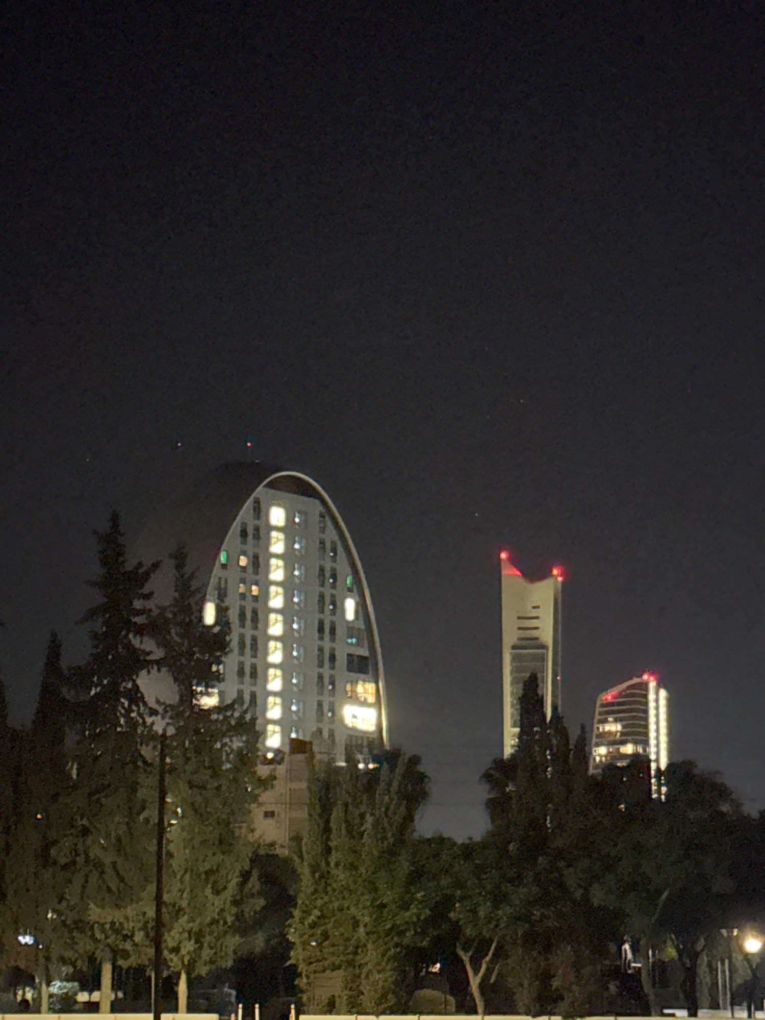 The image captures a nighttime view of the city of Agios Athanasios Municipality, Cyprus. In the foreground, silhouetted trees obscure parts of the scene, with a lamppost visible on the left. In the middle ground, a distinctively shaped, curved white building is illuminated from within, showcasing multiple lit windows. Behind this, a slender, tall skyscraper with red lights at its peak stands prominently. To its right, a shorter, similarly illuminated building with horizontal bands is visible. The overall atmosphere is dark, with the city lights providing the primary illumination.