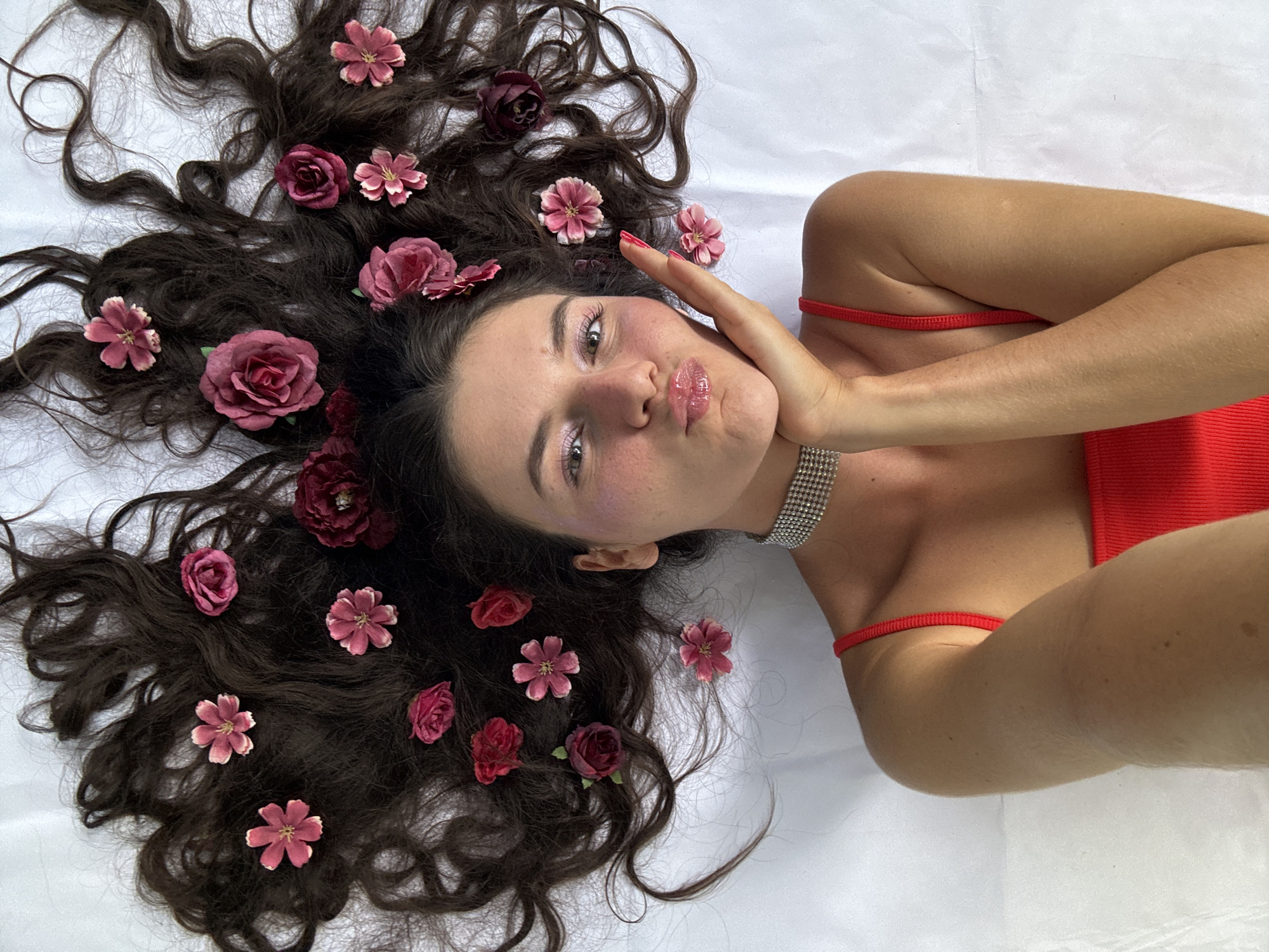 In this media file, a young woman is lying on her back with her head slightly turned towards the camera. Her long, dark, wavy hair is spread out around her head and shoulders, adorned with several artificial flowers in shades of pink and dark red. She is wearing a vibrant red top, possibly a swimsuit or dress, with thin straps. A shimmering, multi-row rhinestone choker is visible around her neck, and a thin red bracelet is on her wrist.
The woman has her left hand resting on her cheek, and her eyes are looking towards the camera. Her lips are pursed, and she appears to be pouting or making a playful expression. Her makeup includes shimmery eyeshadow and a glossy lip color. The overall impression is one of youthful playfulness and a touch of glamour.
The setting is a bright, softly lit environment, suggested by the white background which appears to be fabric, possibly a bedsheet or a studio backdrop. The lighting is even and diffused, casting minimal shadows, which indicates an indoor setting or an overcast day if outdoors. There are no explicit cues in the image to suggest a specific time of day or weather conditions beyond the soft lighting. The location context of Mũi Né, Vietnam, is not visually evident within the image itself, as there are no distinctive landmarks, vegetation, or architectural elements present.
The activity is a posed portrait or selfie, with the woman arranging herself and the flowers for the photograph. The mood is lighthearted and whimsical, enhanced by the floral accents and her playful facial expression. There is no visible text in the media file.