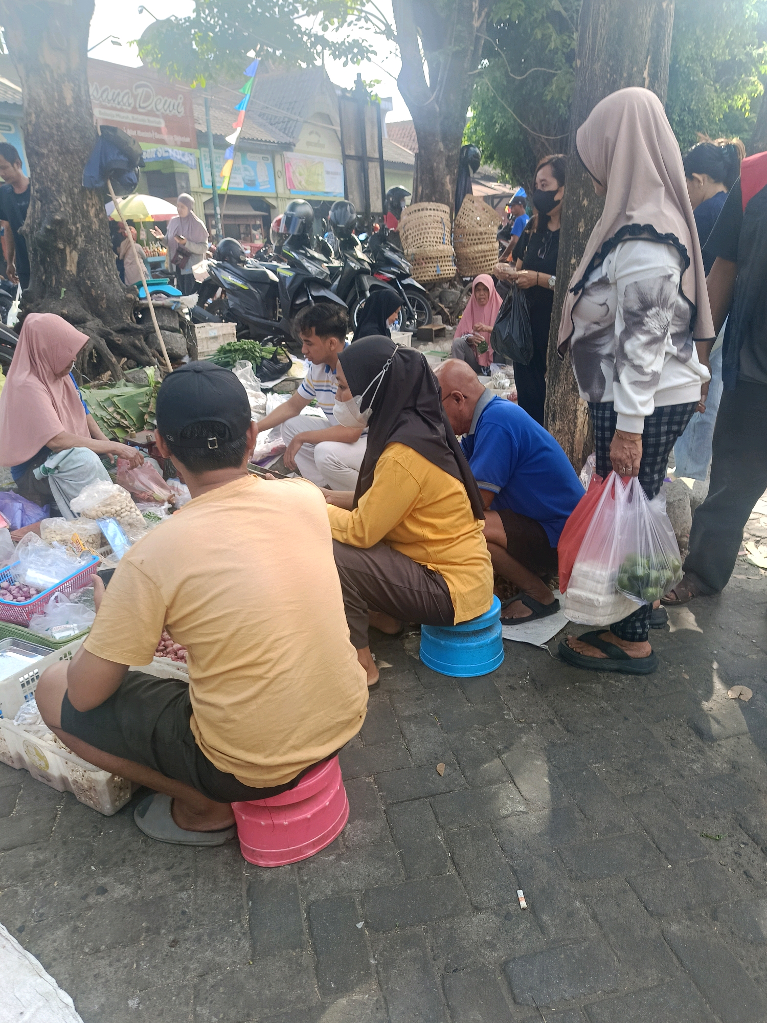 The image depicts a busy outdoor market scene, likely in the city of Yogyakarta, Indonesia, given the context provided by the original prompt. Several individuals are present, some engaged in transactions and others observing. In the foreground, a man wearing an orange t-shirt and a black cap is crouched down, his back to the camera. To his left, a woman in a pink headscarf is seated, seemingly interacting with a basket of goods. To the right of the man in orange, a woman in a yellow top and dark headscarf is also seated, her attention directed towards the ground. Further back, a man in a blue shirt is seated, looking downwards. Other individuals are visible in the background, including a woman wearing a black face mask and holding a plastic bag. Numerous items are displayed for sale, including vegetables, fruits, and other food products, arranged in baskets and bags. Motorcycles are parked in the background, indicating a common mode of transportation. The ground is paved with interlocking bricks, and the scene is shaded by large trees. The overall atmosphere suggests a lively market environment.