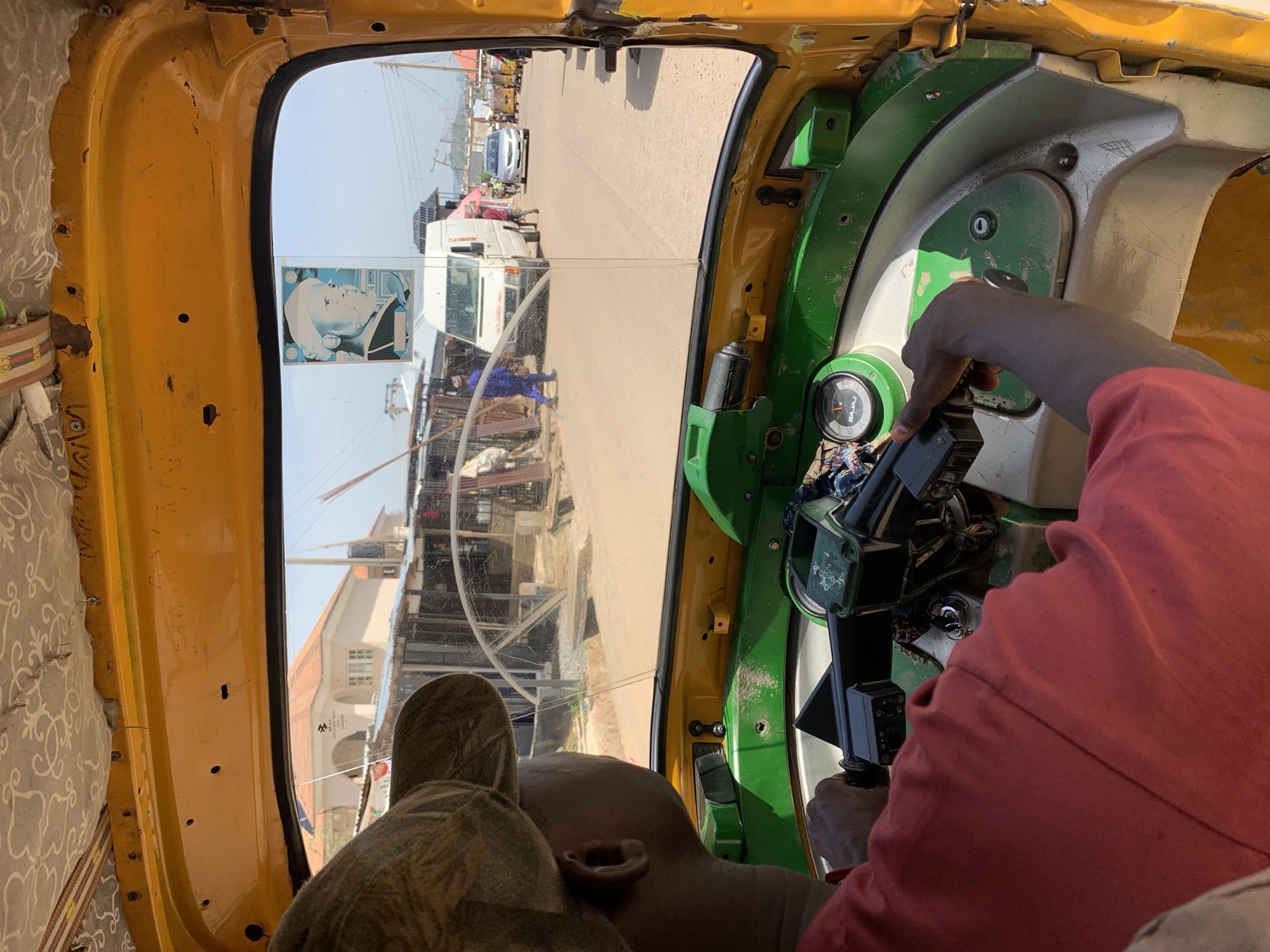 The interior of a yellow and green Bajaj tricycle in Abuja, Nigeria. A dark-skinned individual, wearing a red shirt, steers the vehicle. Through the windshield, a street scene is visible, showing other vehicles and buildings. A white truck is in the distance. A sticker of a man's face is on the window. The vehicle is moving on a sunny day.
