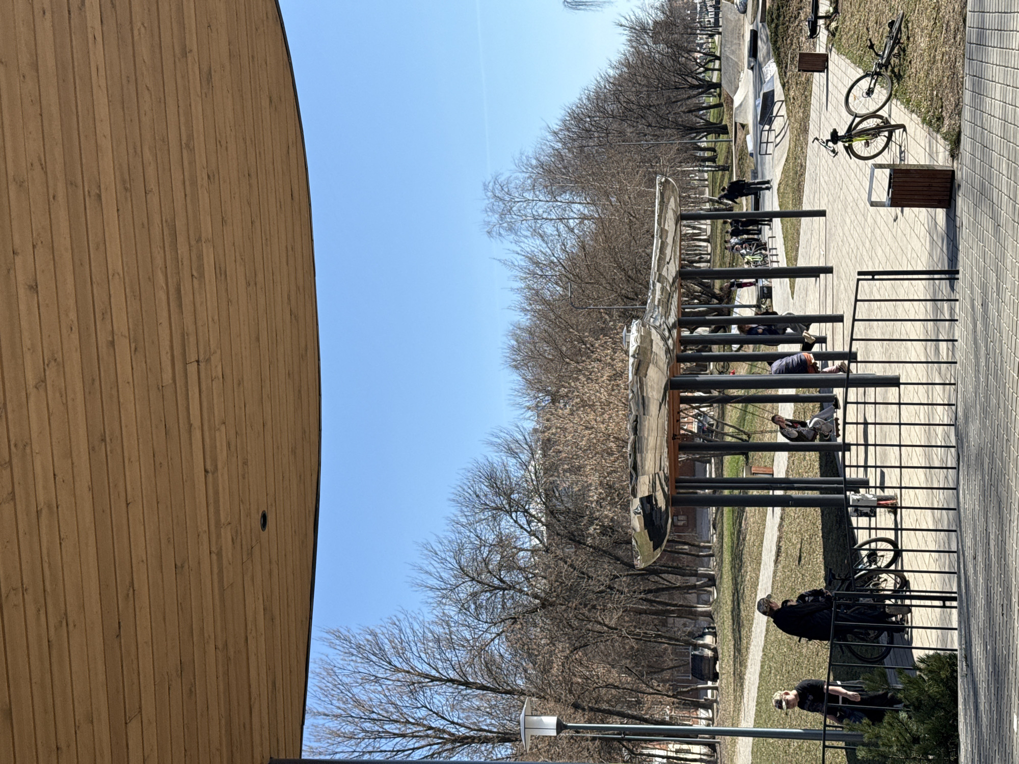This image captures an outdoor scene, likely a public park or recreational area, on a clear and sunny day. The season appears to be late winter or early spring, as most trees visible in the background and mid-ground are bare.
On the left, a large, curving wall clad in vertical wooden slats forms a prominent architectural feature. The sky above is a bright, clear blue.
In the center of the frame, there is a modern outdoor fitness or playground structure made of reflective metal, featuring multiple vertical poles and horizontal bars. Several people are engaged with this equipment; one person is lying down on a lower bar, and another is seated on a higher section.
A paved pathway, composed of light grey square tiles, extends through the right side of the image, alongside grassy areas. Two bicycles are parked near the path. Two individuals are standing close to the bikes, one wearing a dark jacket and cap, and another in a black t-shirt and light-colored headwear. Further in the background, more people are scattered across the park area. Metal railings and fences are also visible, delineating parts of the space.
The overall impression is one of casual outdoor activity and relaxation under pleasant weather conditions. There is no visible text.