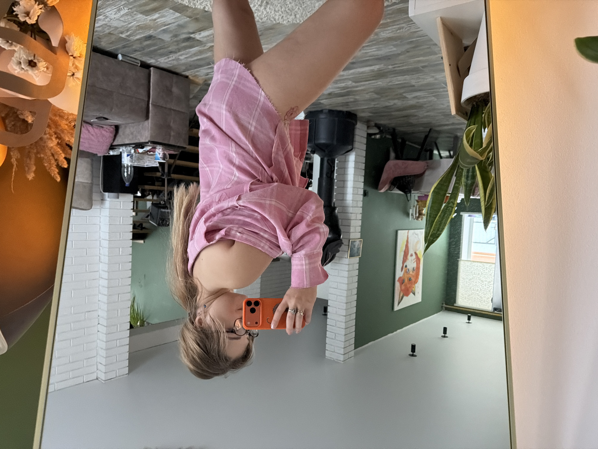 A woman is seen reflected in a mirror indoors. The mirror captures a portion of a living room with a couch, a painting on the wall, and various decorations. The woman is holding an orange phone and appears to be taking a picture or video of herself in the mirror. The location is indoors and likely in a residential setting, potentially in the city of Большая Шильна, Russia.