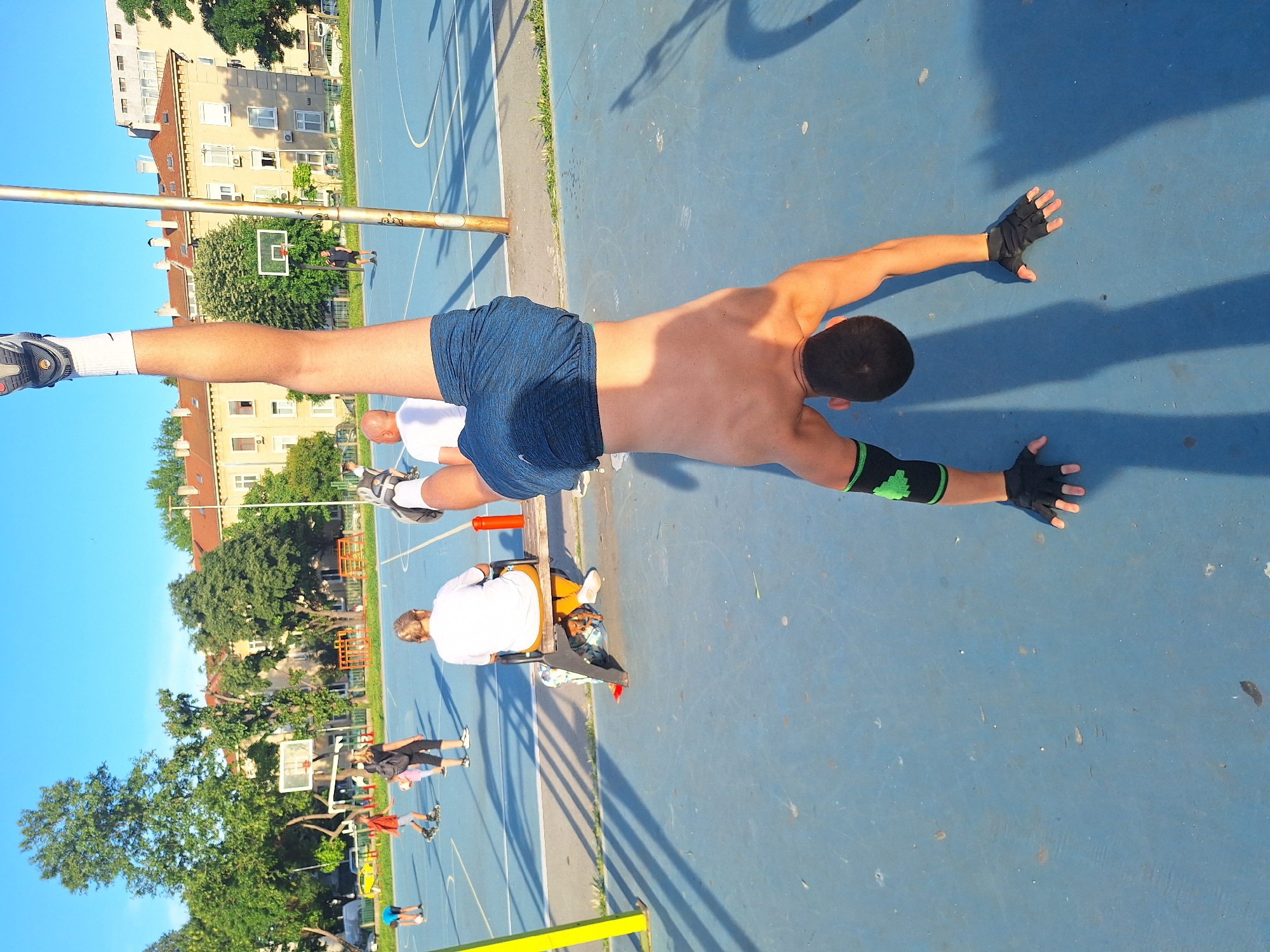 An individual is performing a handstand on a blue outdoor basketball court. The person is positioned in the foreground, with their legs extended upwards and their body inverted. They are wearing blue shorts, black fingerless gloves, and neon green wristbands. In the background, several other people are visible on the court, some playing basketball and others sitting on a bench. There are basketball hoops and net structures around the court. Trees and apartment buildings are visible beyond the court, under a clear blue sky. The scene appears to be in a city park or recreational area. The overall lighting suggests it is daytime, with long shadows cast by the people and objects.