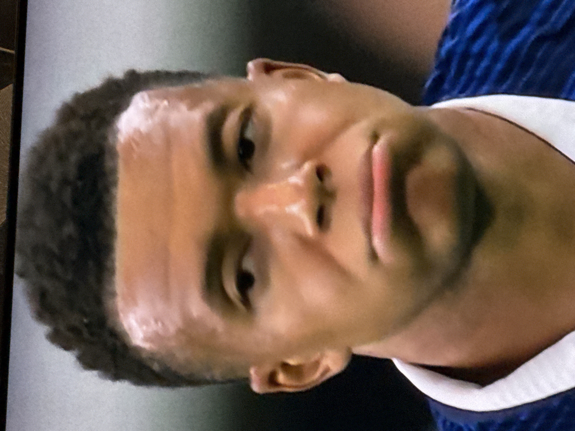 The image shows a close-up, head-and-shoulders view of a young Black man, likely a soccer player, wearing a blue and white jersey. He has short, dark, curly hair and a clean-shaven face. His expression is pensive, with his mouth slightly downturned and his eyes looking to the left. The lighting suggests he is indoors, possibly on a field under stadium lights, with a blurred background. There is no indication of the city of Malmyzh, Russia, or any other specific location within the image.