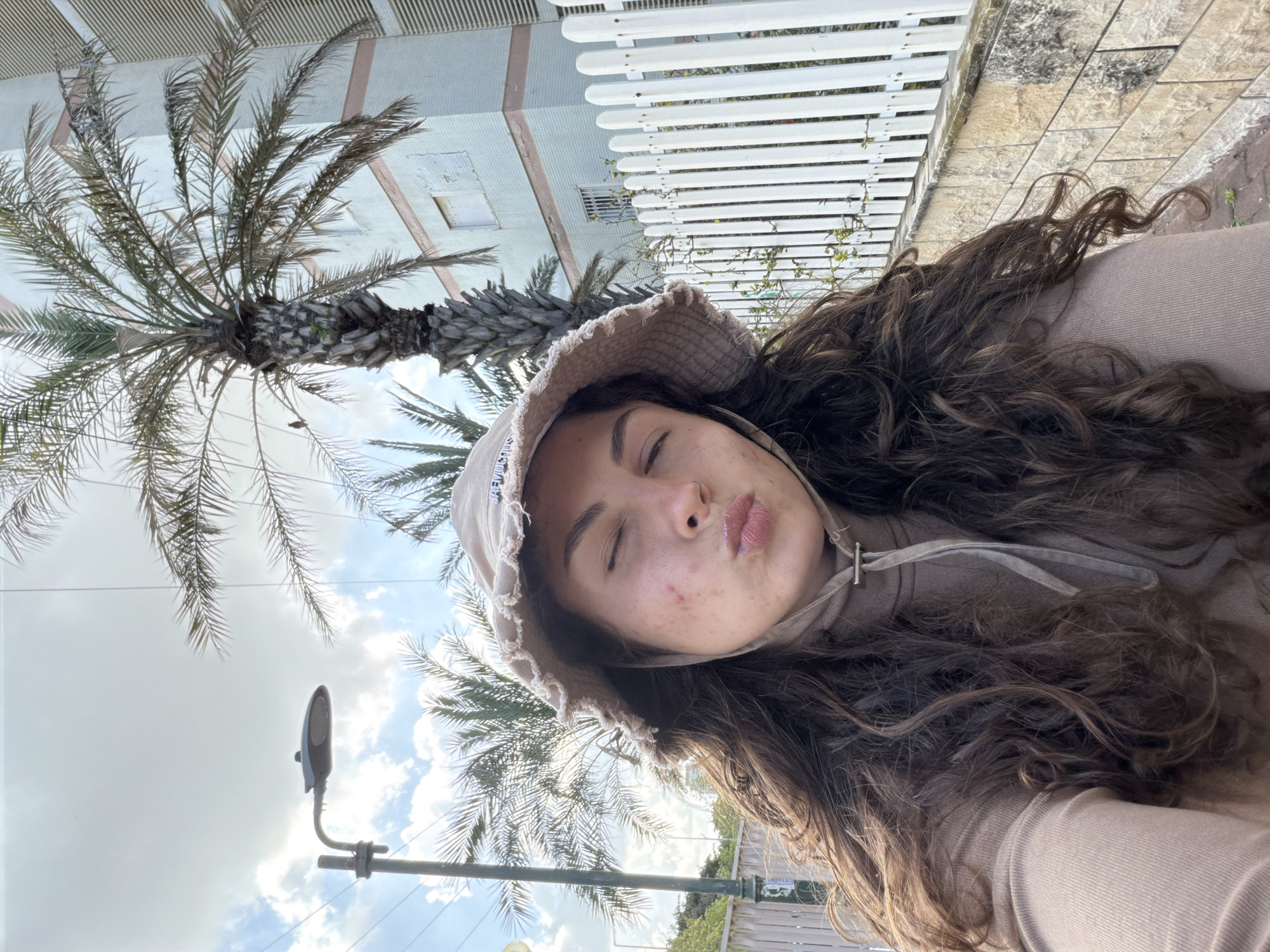 A woman with long, dark brown curly hair is taking a selfie outdoors. She is wearing a light brown hoodie and a matching bucket hat. She has her eyes closed and her lips pursed. Behind her, there's a palm tree, a street lamp, and a building with a white picket fence. The photo was taken in Kiryat Bialik, Israel.