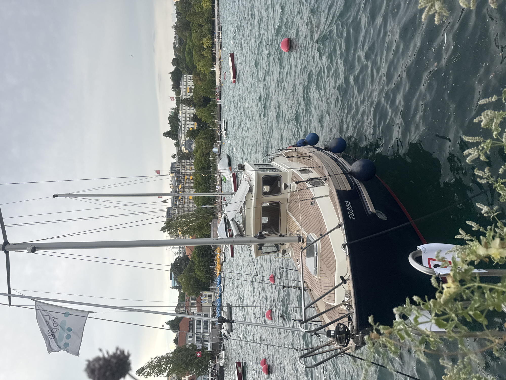 This is a medium shot, eye-level, outdoor, daytime, vertical photograph of a sailboat docked in a harbor on Lake Geneva, Lausanne, Switzerland. The boat, with the registration "VD2013" visible, is dark blue with a light-colored wooden deck and a white cabin. It is equipped with a mast, rigging, and several blue fenders. Red buoys are scattered in the water around the boat. In the background, across the lake, a city skyline with numerous buildings, trees, and flags is visible. Two Swiss flags are prominent on buildings. The sky is overcast. There are no visible people interacting with the boat.
