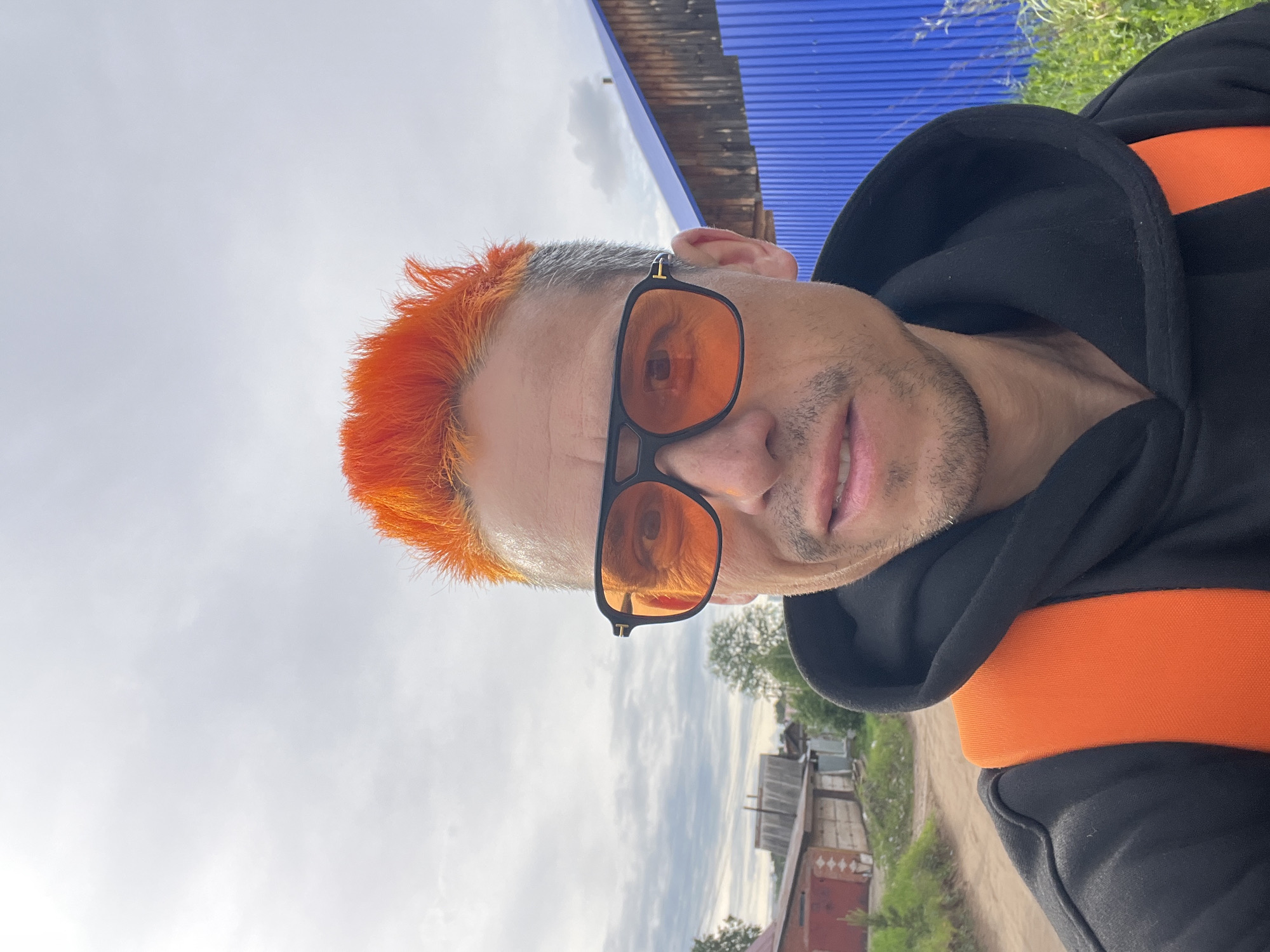 A man with orange hair and sunglasses is shown in a close-up selfie. He is wearing a black hooded sweatshirt with an orange stripe across the shoulders. The background features a cloudy sky, a blue corrugated metal roof, a glimpse of a rural street, and houses. It is not possible to verify the location of Chaikovsky, Russia.