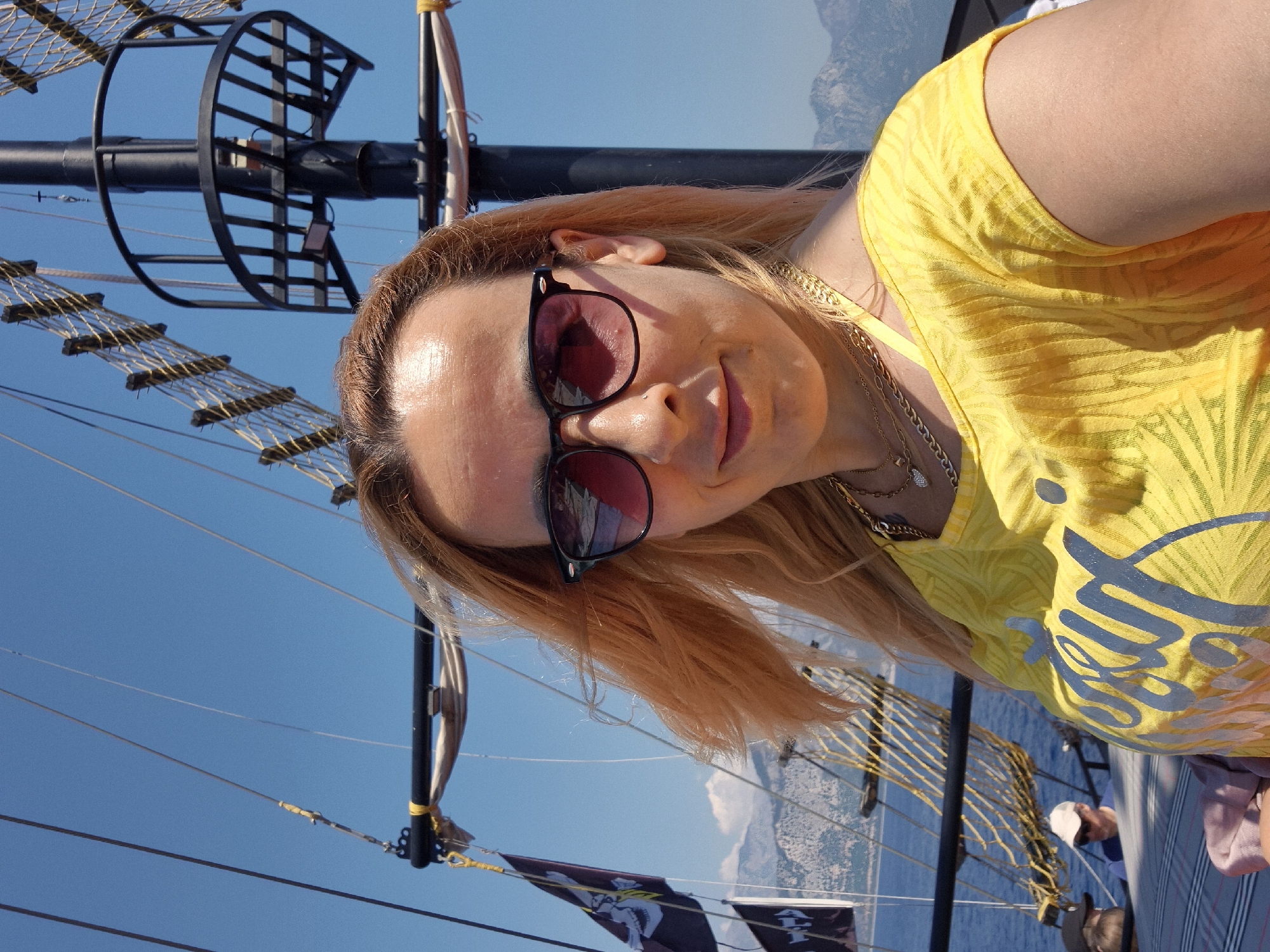 This is a slightly rotated, close-up selfie of a woman on a boat. She is wearing sunglasses and a bright yellow, textured t-shirt with a palm leaf pattern and some grey text on the front. She has blonde hair and is wearing necklaces. The background shows a clear blue sky, rigging and part of a mast from the boat, and a distant, hazy coastline. The sunlight suggests it is daytime, likely a sunny day. The woman appears to be smiling slightly, her expression is relaxed and happy. The scene suggests a leisure activity, possibly a boat trip or vacation. There is some visible text on her shirt, but it is stylized and difficult to read clearly in this image.