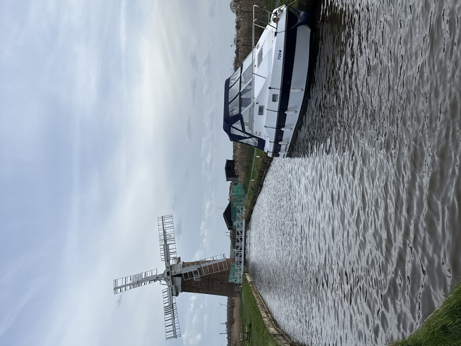 This image depicts Polkey's Mill, a brick tower windmill, located within the Norfolk Broads area of North Norfolk, United Kingdom. The mill, with its four-arm skeleton sails, stands prominently on the left bank of a narrow, rippled waterway, likely a canal or river. A white metal footbridge or walkway extends across the waterway from the mill's base. To the right, a white and dark blue motor cruiser named "Moon Dancer," bearing the registration "706W," is moored parallel to the right bank. The boat has a blue canvas cover over its cockpit area and is secured with white fenders. The banks of the waterway are covered in green grass, with a wooden bench visible on the left bank near the foreground. Beyond the mill, small, dark-colored sheds and areas of tall, dry reeds are visible. The sky is uniformly overcast with light grey clouds. No individuals are present in the scene.