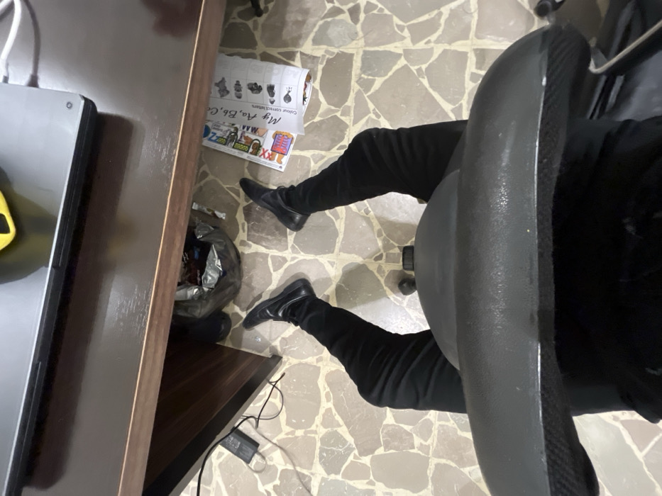 A person sits in a gray office chair, clad in black pants and shoes, with their legs stretched forward. A desk is partially visible on the left, holding a laptop. A plastic bag, filled with items, rests beside the desk. On the floor, a children's alphabet poster lies near the legs. The floor is tiled with irregular beige shapes. Cables run from under the desk.