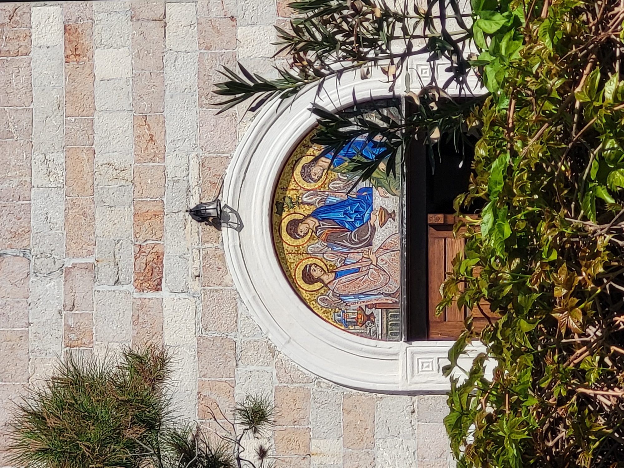 The image shows the exterior of a building in Budva, Montenegro. The wall is constructed of rectangular stone blocks, alternating between lighter and darker shades. A circular window is present, featuring a mosaic depicting three religious figures with golden halos, rendered in blue, brown, and gold hues. The window is framed with a white border. A black, ornate light fixture is attached to the wall near the window. Green foliage partially obscures the window and the surrounding wall. A smaller, different type of green plant is at the bottom of the wall. An open, dark wood shutter is visible to the right of the mosaic.