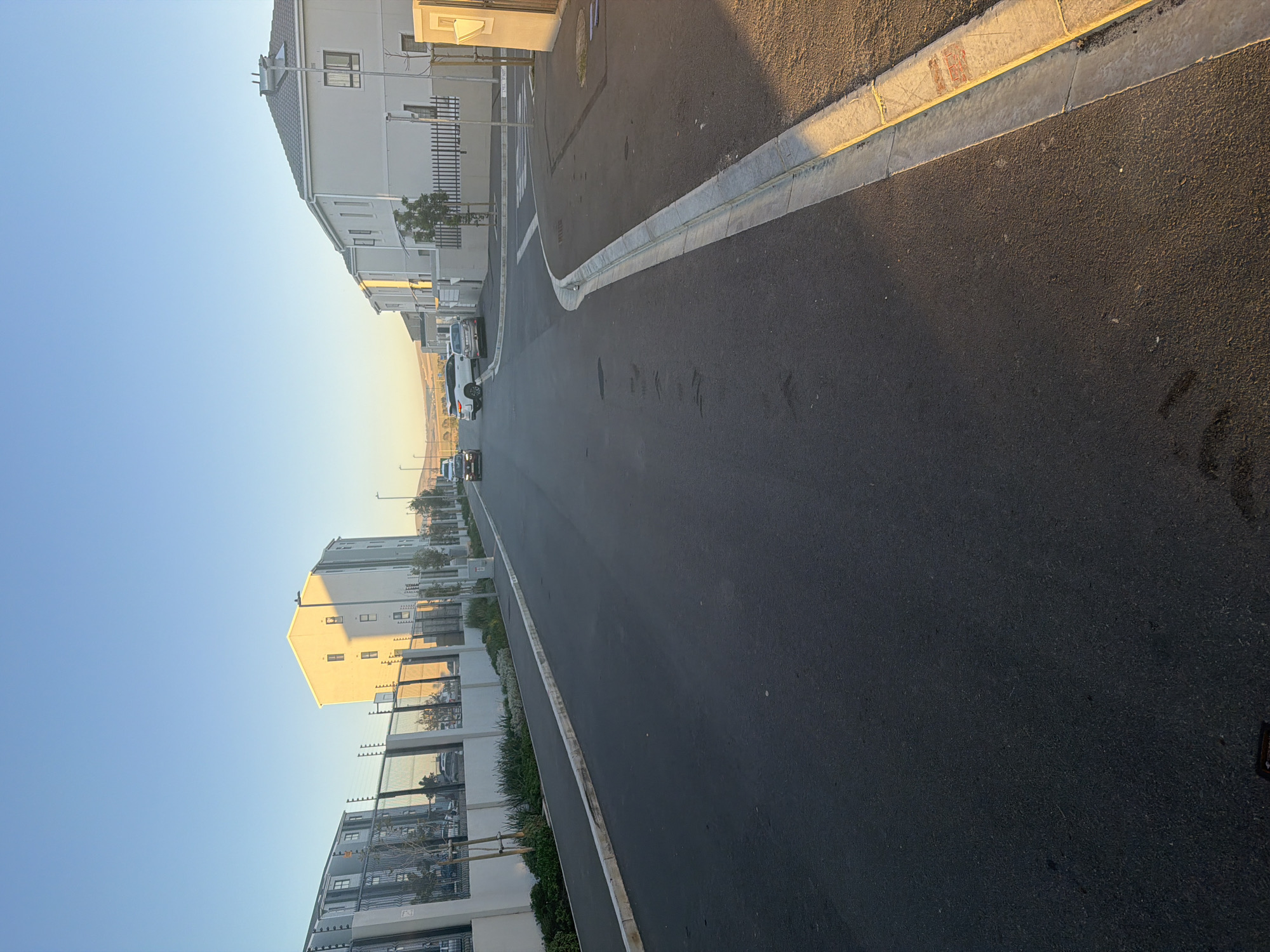 The media file displays an outdoor street scene in what appears to be a modern residential or mixed-use development, likely during either sunrise or sunset. The image is captured from a low angle, looking down a gently curving asphalt road that recedes into the distance.
The street itself is dark asphalt, appearing well-maintained. It is bordered by concrete curbs and gutters on both sides. On the right side, the curb has areas of yellowing or discolored paint. The road shows no significant markings in the foreground but there are faint indications of white lines further down.
Modern, multi-story buildings line both sides of the street. On the right, the buildings are predominantly light-colored (white, beige, or light grey) with multiple windows and some with visible balconies or rooftop structures. One prominent building on the right foreground features a gabled roof and is strongly illuminated by warm, golden sunlight. On the left side of the street, there are also multi-story buildings, some with metal fences or railings along their perimeters. The architecture suggests relatively new construction.
Several vehicles are parked along the left side of the street, including a white sedan and a darker vehicle. More vehicles are visible further down the street on both sides. Small trees and various shrubs are planted along the sidewalks and fences, adding greenery to the urban environment.
The sky is a clear, bright blue, fading slightly to a lighter hue near the horizon. The sun is low in the sky, casting long shadows across the street and highlighting the facades of the buildings it directly illuminates, creating a strong contrast between light and shadow. This warm lighting strongly suggests either early morning or late afternoon.
No people are visible, and there is no direct activity taking place, conveying a sense of quietude. No distinct, legible text is visible within the image. The scene is consistent with a newly developed area in Cape Town, South Africa, as per the provided context.