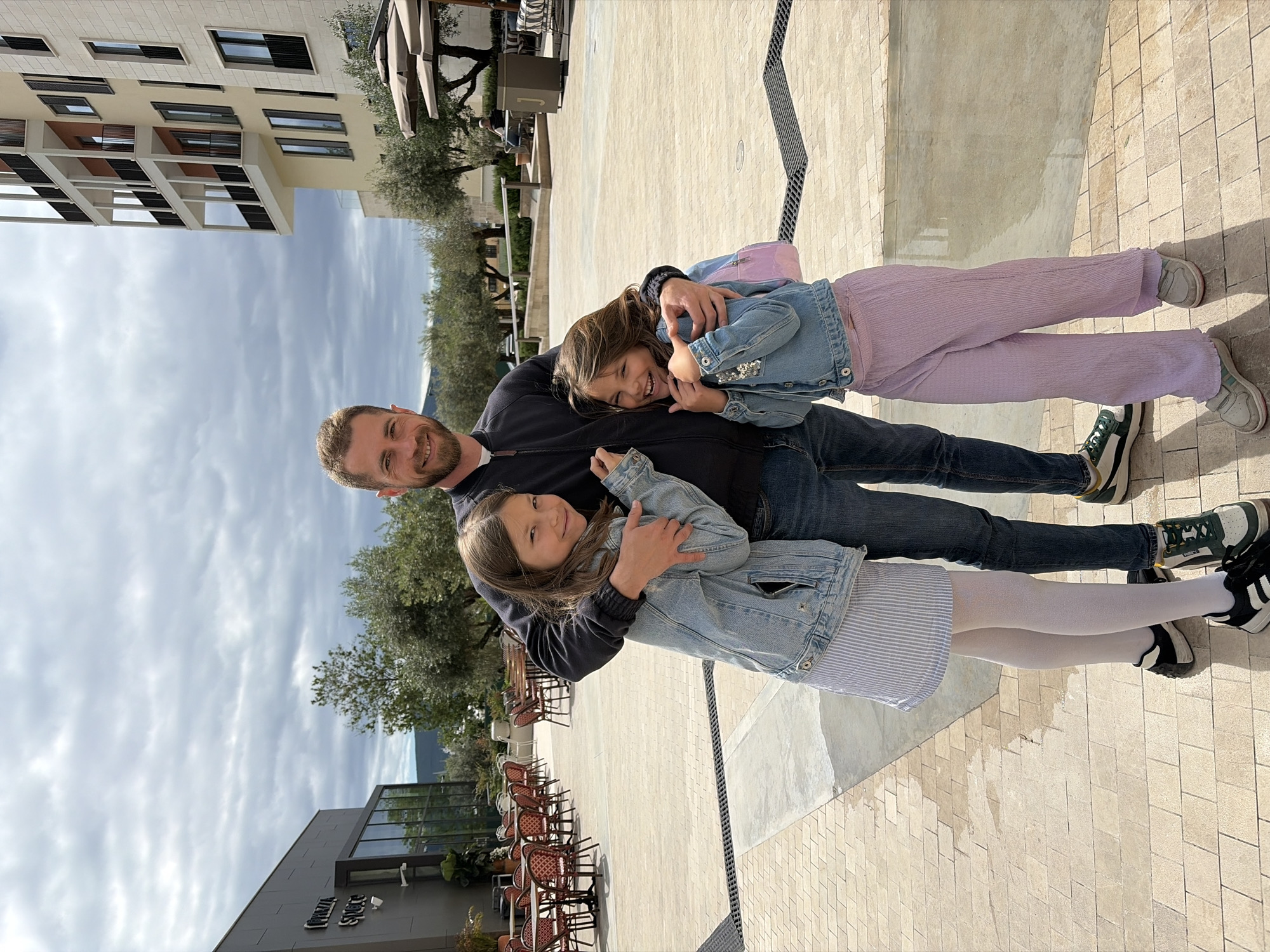 A man is posing with two young girls in Tivat, Montenegro. The man has short, light brown hair and is wearing a dark jacket and jeans. The two girls are hugging him and smiling. One is wearing a denim jacket, striped dress, and white tights with black and white sneakers. The other is wearing a denim jacket and light purple pants with sneakers. They are standing on a light-colored bricked surface. In the background are buildings, trees, and outdoor furniture, like tables and chairs, indicating a restaurant or cafe area. The sky is overcast.