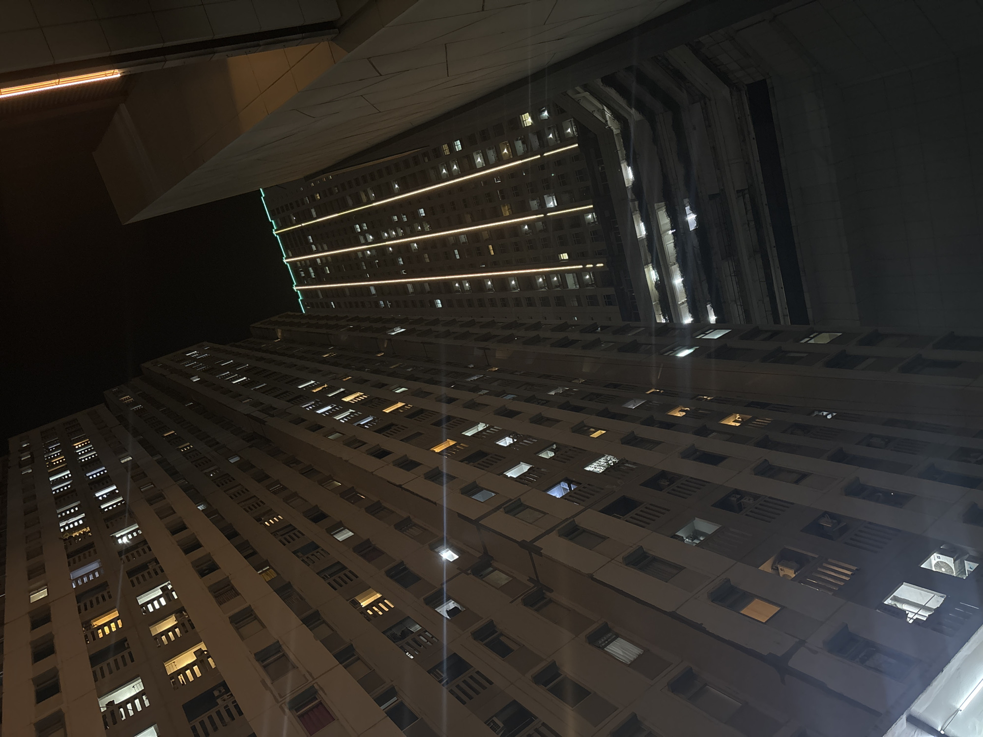 The image depicts a night view of a residential building in North Jakarta, Indonesia. The perspective is looking upwards at the facade of the building, which is illuminated by various lights from within its numerous windows. The building is tall and appears to be made of concrete. The windows are rectangular and some have balconies with railings. Several air conditioning units are visible on the balconies. Some windows are dark, indicating unoccupied apartments, while others glow with warm yellow or white light, suggesting residents are present.
To the right of the main residential building, another structure is visible, made of a lighter colored material and having a more angular design. This structure is also illuminated by some lights, particularly a vertical strip of warm orange light running along its edge.
In the foreground, a portion of a darker, possibly more modern building is seen, with a slanted geometric facade. The sky above is dark, indicating it is nighttime. The overall scene captures the atmosphere of a densely populated urban environment at night, with the lights of the buildings creating a stark contrast against the dark sky. The image is taken at a slight angle, giving a dynamic feel to the composition.