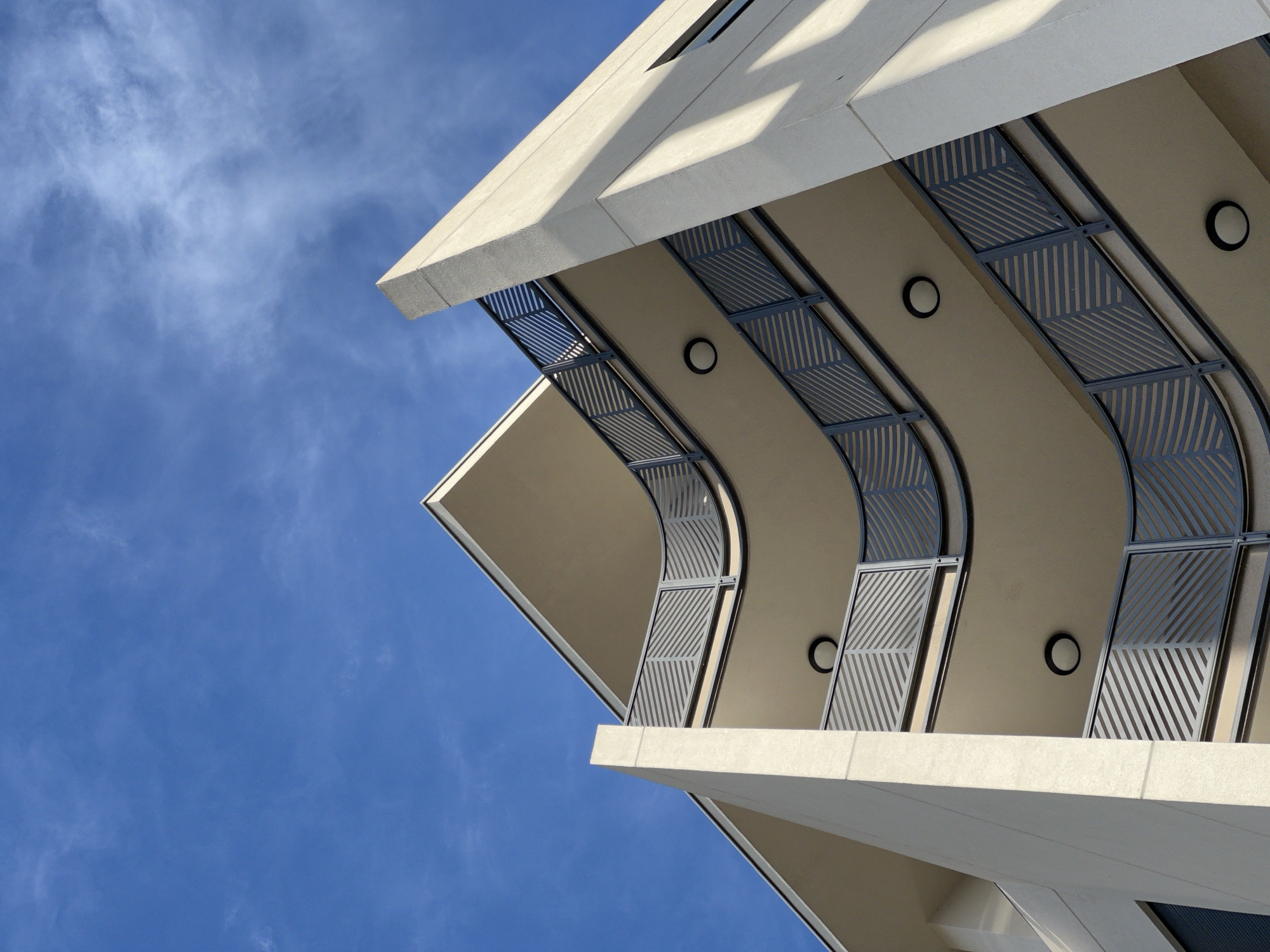 A low-angle shot of a modern building against a blue sky with faint clouds. The building is angular with beige-colored walls and white concrete edges. The building's balconies feature curved metal screens with linear patterns, punctuated by circular lights. The image is focused on the architectural details and the contrast between the building and the sky. Without additional context, pinpointing the precise location within Bar, Montenegro is not possible.