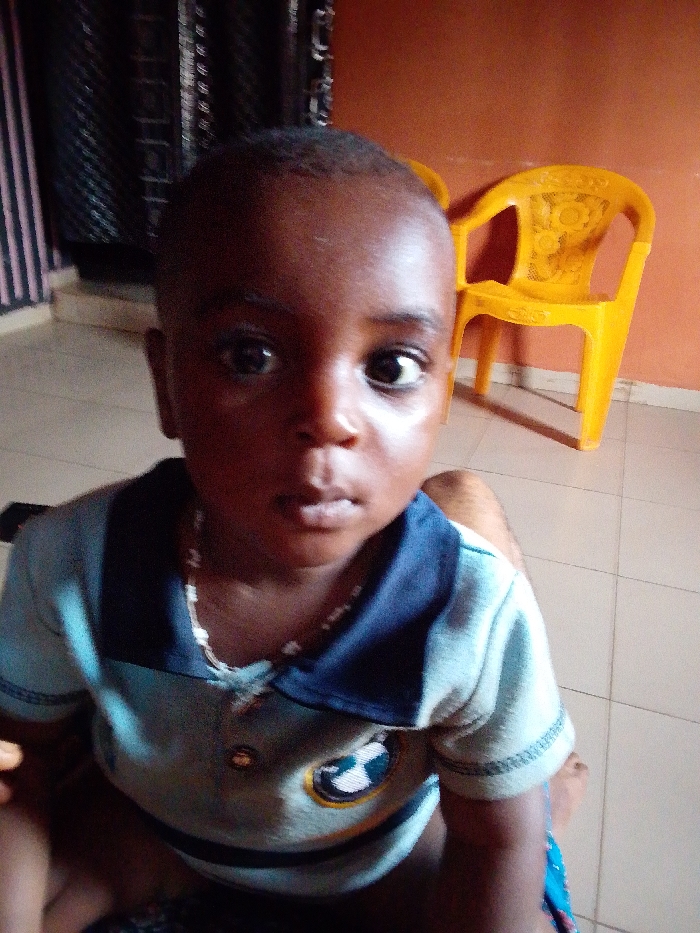 An eye-level, close-up indoor shot features a young child, likely a toddler or infant, with dark skin and closely cropped dark hair. The child is positioned centrally in the frame, looking directly into the camera with wide, dark eyes and a neutral, slightly serious expression. They are wearing a light blue polo shirt with a dark blue collar and what appears to be horizontal stripes or color-blocking. A small, circular emblem or logo is visible on the left side of the shirt (from the viewer's perspective), featuring a white and dark checkered pattern within a golden or yellow ring. A light-colored, possibly white or cream, beaded necklace is worn around the child's neck.
The child appears to be seated, with a portion of what looks like an adult's bare knee or leg visible in the lower right corner, suggesting the child might be on someone's lap or very close to an adult.
The background is split into two main sections. To the left of the child, a dark, vertically striped curtain or fabric is visible, possibly black with lighter vertical lines. To the right, behind the child's head, there is a plain, solid orange or terracotta-colored wall. Against this wall, a bright yellow plastic chair with an intricately patterned backrest (featuring what looks like a floral or sunburst design) stands out. The floor beneath the child and the chair is made of light-colored, possibly beige or off-white, square tiles. The lighting appears to be ambient indoor light, casting some gentle shadows and highlights on the child's face. No text is visible in the image.