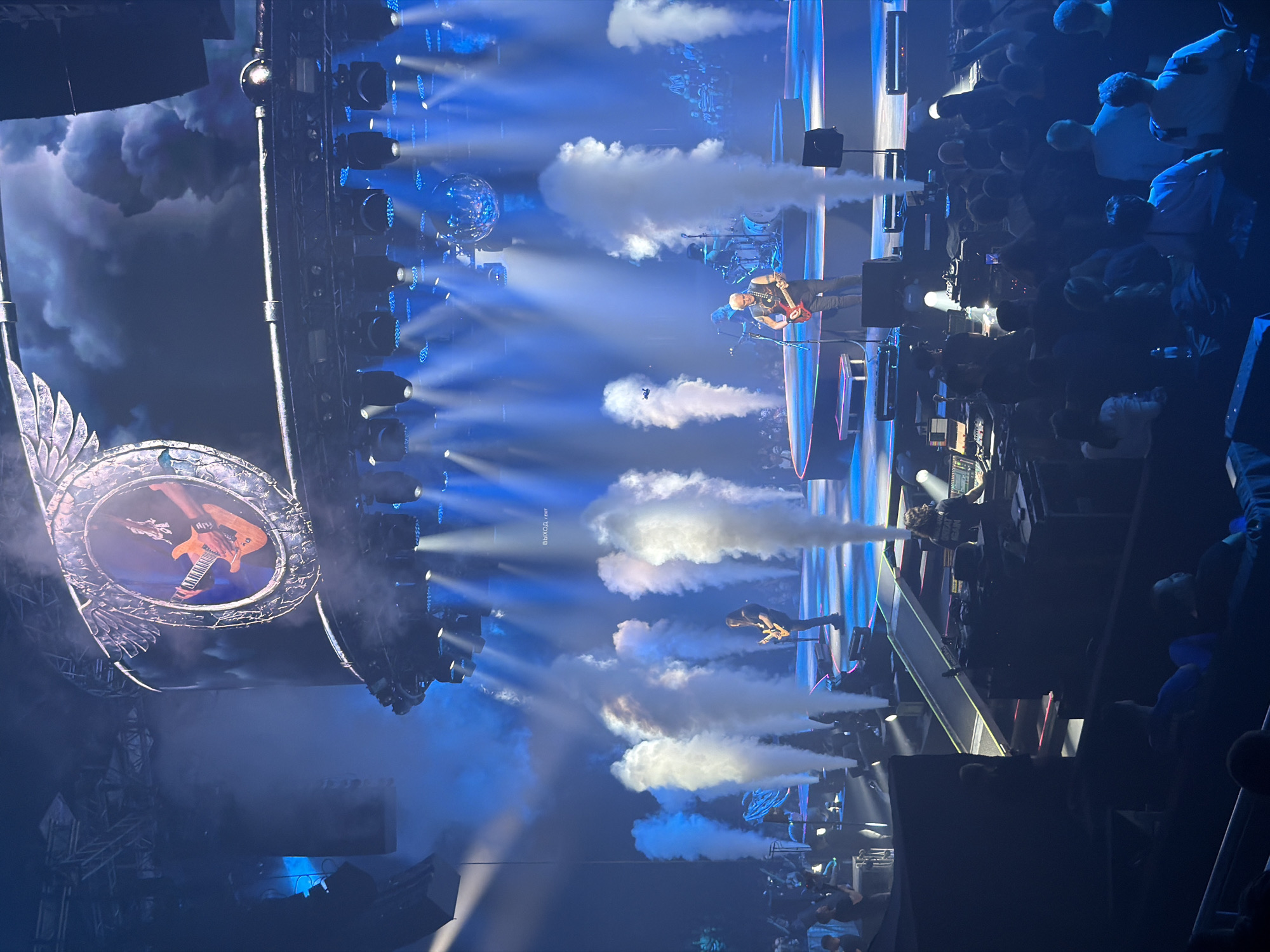 The image captures a live concert performance with a dramatic stage setup. The stage is illuminated with bright blue spotlights, and plumes of white smoke are strategically released, creating a dynamic and atmospheric effect. On stage, several musicians are performing. One guitarist, wearing a black t-shirt and jeans, is prominently positioned in the foreground, playing a red electric guitar. Behind him, another musician can be seen playing drums. A large screen is suspended above the stage, displaying an image of a hand playing a guitar within a circular frame, framed by winged elements. The audience is visible in the lower section of the image, seated in a darkened venue, facing the stage. The overall impression is of a high-energy rock concert. There is no specific indication or visual information within the image to suggest the location being the city of Saint Petersburg, Russia.