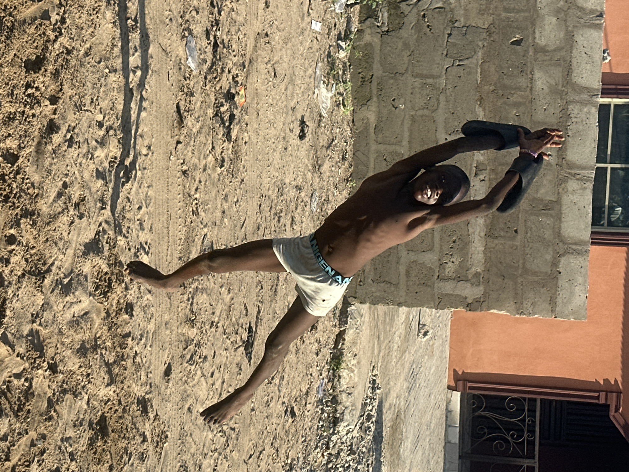 A young boy, likely in his early teens, is captured mid-jump in this photograph. He is shirtless and wearing white boxer briefs with a blue waistband. His body is arched backward, and he is holding a pair of black flip-flops above his head with both hands. His facial expression is one of exertion and perhaps joy. The ground beneath him is sandy and uneven, with some debris and tire tracks visible. To his right, a rough-hewn concrete wall forms the side of a building, painted in places with an orange hue. A window with a grid is visible on the building. The lighting suggests it is daytime, with strong shadows indicating the sun is relatively high. The overall scene depicts a moment of energetic play in an outdoor, somewhat rustic setting, likely a street or yard in Badagry, Nigeria.
