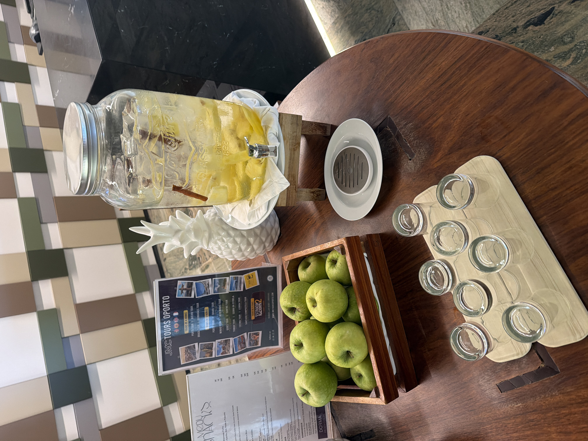 The image depicts a self-service beverage station, likely within a hotel or restaurant. A large glass dispenser filled with ice, lemon slices, and cinnamon sticks, contains a clear liquid, presumably infused water. It is positioned on a wooden stand. Beside it, a white decorative pineapple and a brochure for "JMC TOURS OPORTO" are visible. In the foreground, a wooden crate overflows with fresh green apples. A tray with six small shot glasses is placed on a polished wooden table. The table's surface exhibits wood grain and a decorative inlaid pattern. In the background, a wall with a geometric pattern of squares in beige, green, and white tones is visible. A dark countertop is also present to the right. The scene appears to be set in a contemporary establishment, possibly in Espinho, Portugal, given the tour brochure. No people are present in the image.