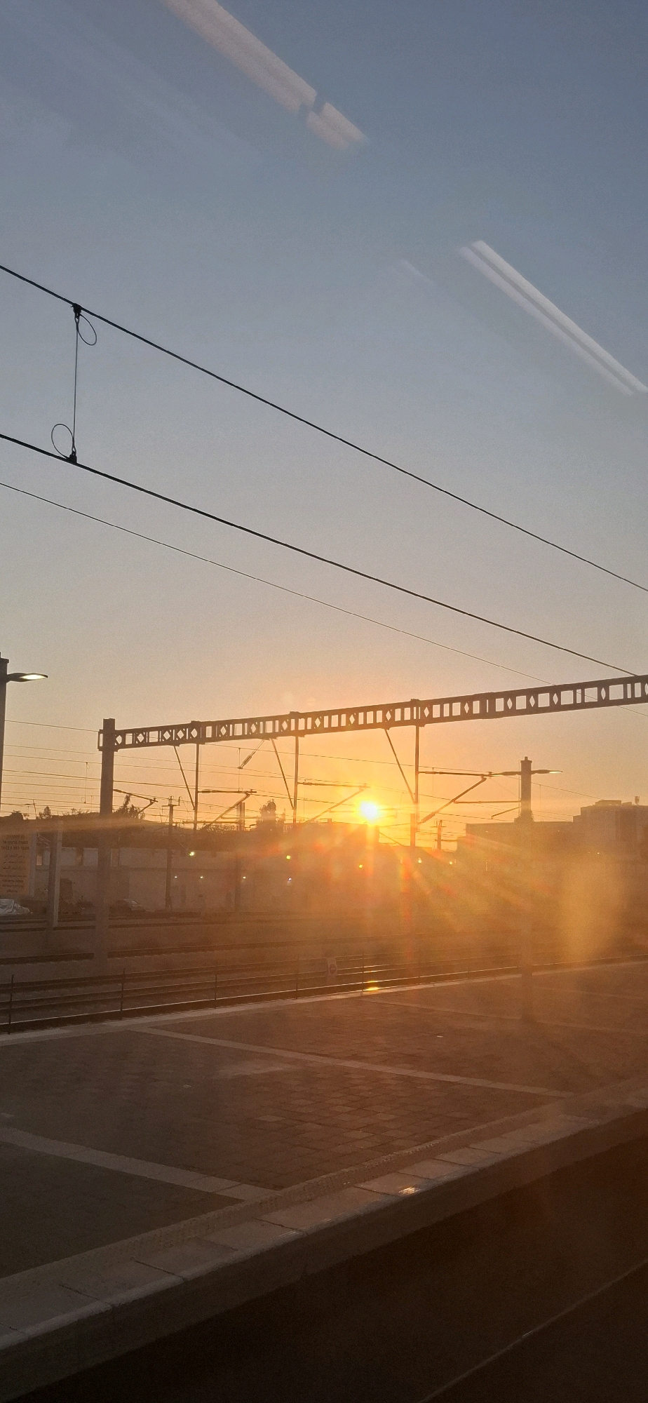 The image depicts a sunset at a train station in Be'er-Sheva, Israel. The sun, a bright orb, is positioned near the horizon, casting a warm, golden glow across the scene. Lens flare effects are visible, suggesting the sun's intense light. Overhead, a network of electrical wires for the trains crisscrosses the sky, suspended from poles and a decorative lattice structure. In the foreground, the tiled platform of the train station is visible, with tracks running parallel to it. The background shows buildings and structures typical of an urban environment, silhouetted against the colorful sky. The overall impression is one of a serene, late-day atmosphere at a transportation hub.