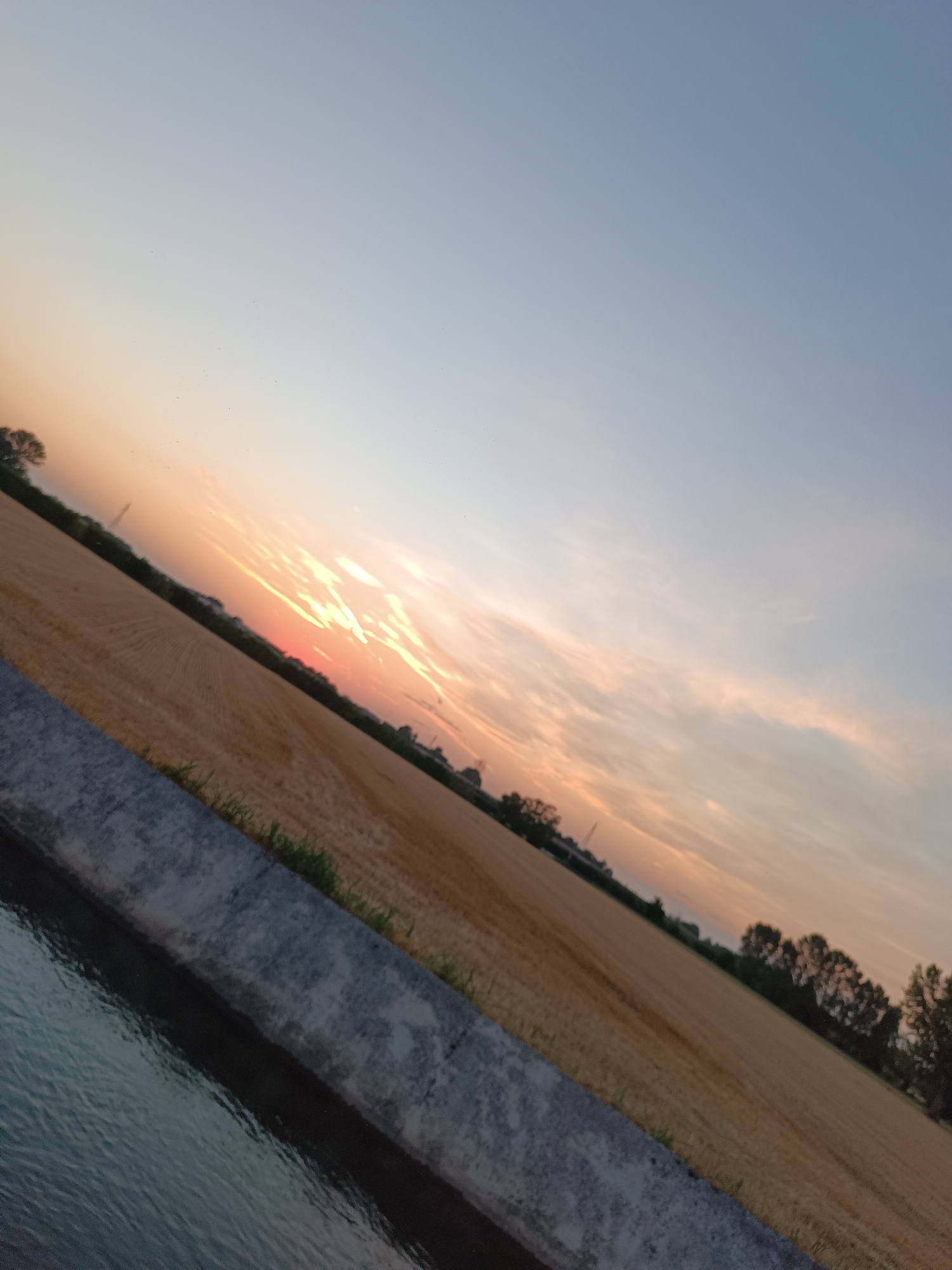 The image depicts a sunset over a rural landscape, likely in the vicinity of Viadana, Italy, given the provided context. The sky is a gradient of pale blue and soft orange, with wispy clouds tinged by the setting sun's light. In the foreground, a concrete channel runs diagonally across the frame, filled with dark, rippling water that reflects the sky's colors. Adjacent to the channel is a field of dry, golden crops, possibly wheat or hay, showing evidence of harvesting with visible rows. A line of trees and distant structures, including what appears to be a utility pole, are silhouetted against the horizon. The overall impression is one of tranquility and the end of a day in a rural setting. There are no people or animals visible in the scene.