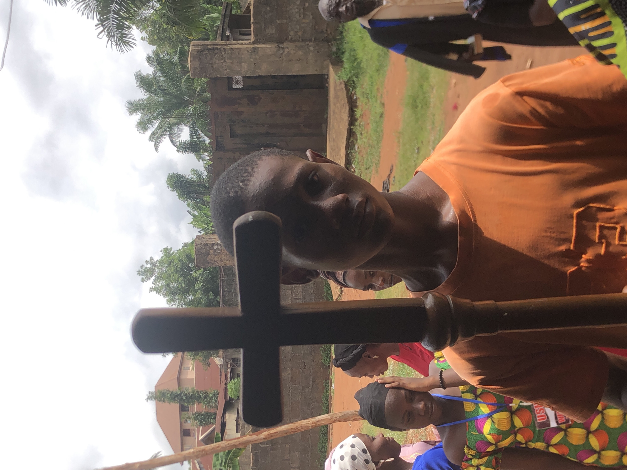 The image depicts a gathering in Ukwu-Abwa, Nigeria, where a man in an orange shirt holds a wooden cross in the foreground. The background shows a group of people; some have hands placed on their heads. Buildings and trees are visible in the surroundings. The overall setting suggests a community event or religious gathering.