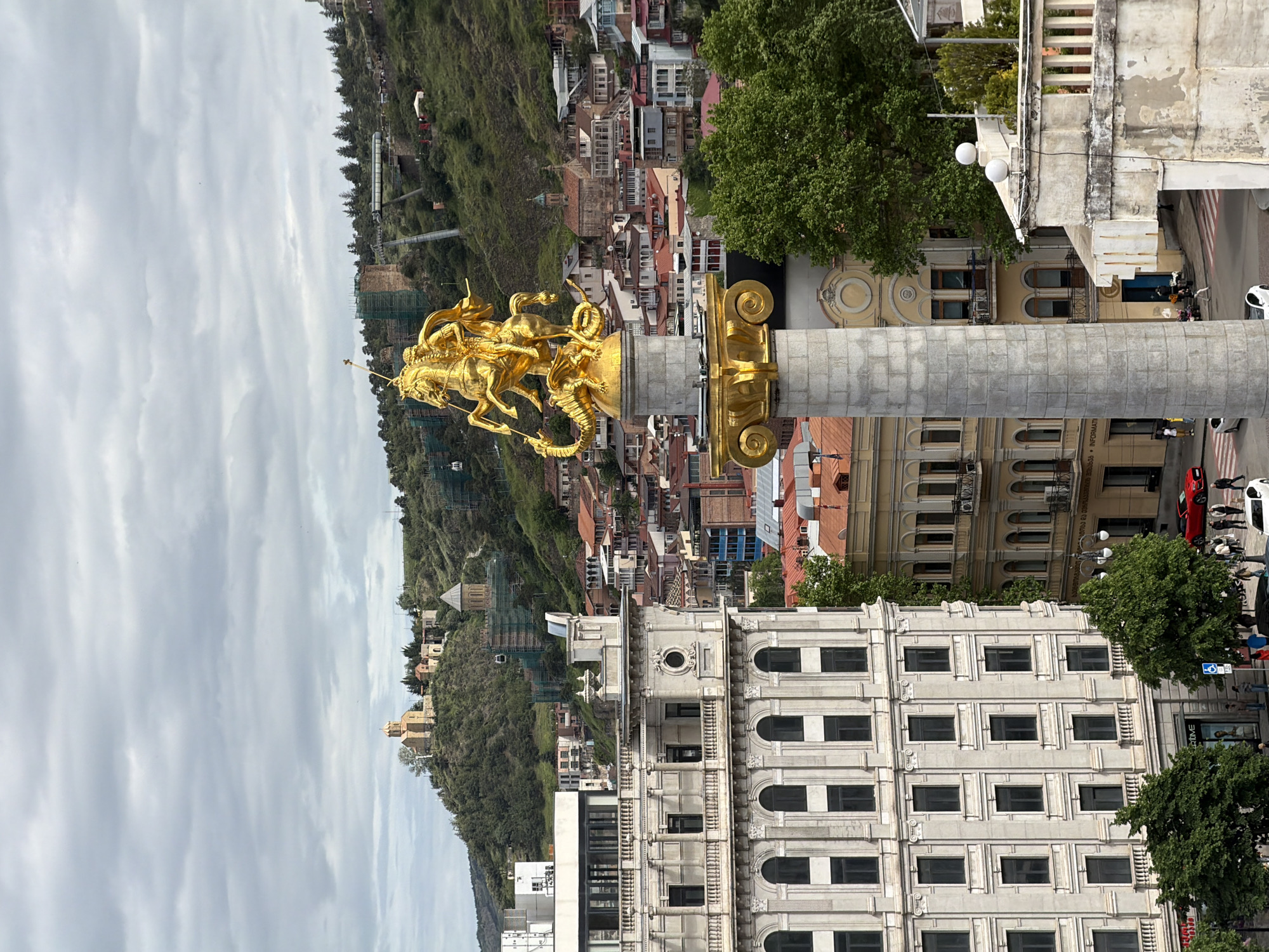The image depicts a view of Tbilisi, Georgia, dominated by the King Vakhtang Gorgasali monument. The monument, topped with a golden sculpture of the king on horseback slaying a dragon, stands prominently in the foreground. A cylindrical stone column with golden Corinthian-style decorations supports the statue. In the background, a dense urban landscape of red-roofed buildings and streets stretches out towards tree-covered hills and a cloudy sky. An off-white building with evenly spaced windows is below the monument. Pedestrians and cars can be seen on the streets.