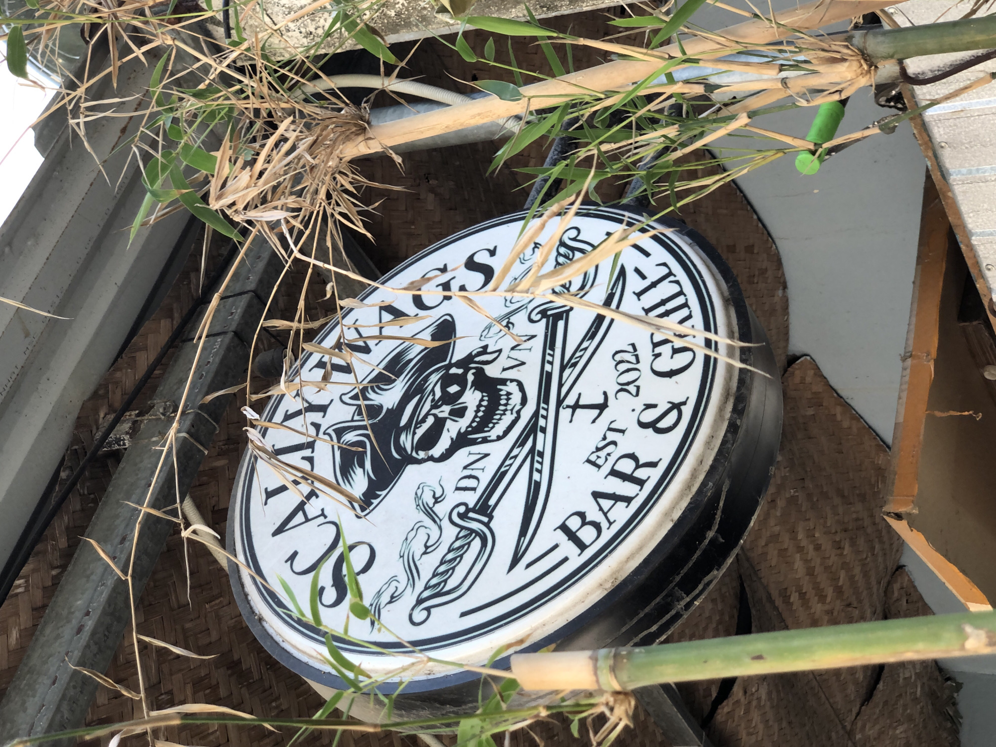 A circular sign for "SCALLYWAGS BAR & GRILL" is partially obscured by bamboo stalks. The sign features a pirate skull wearing a bandana and hat, crossed cutlasses, and text indicating "EST. 2022" and "DN VN". The sign is mounted on a textured, woven background, possibly a ceiling or wall. The environment appears to be outdoors, with natural light. The bamboo is a mix of green and dry stalks. The sign is black and white with some dark outlines.