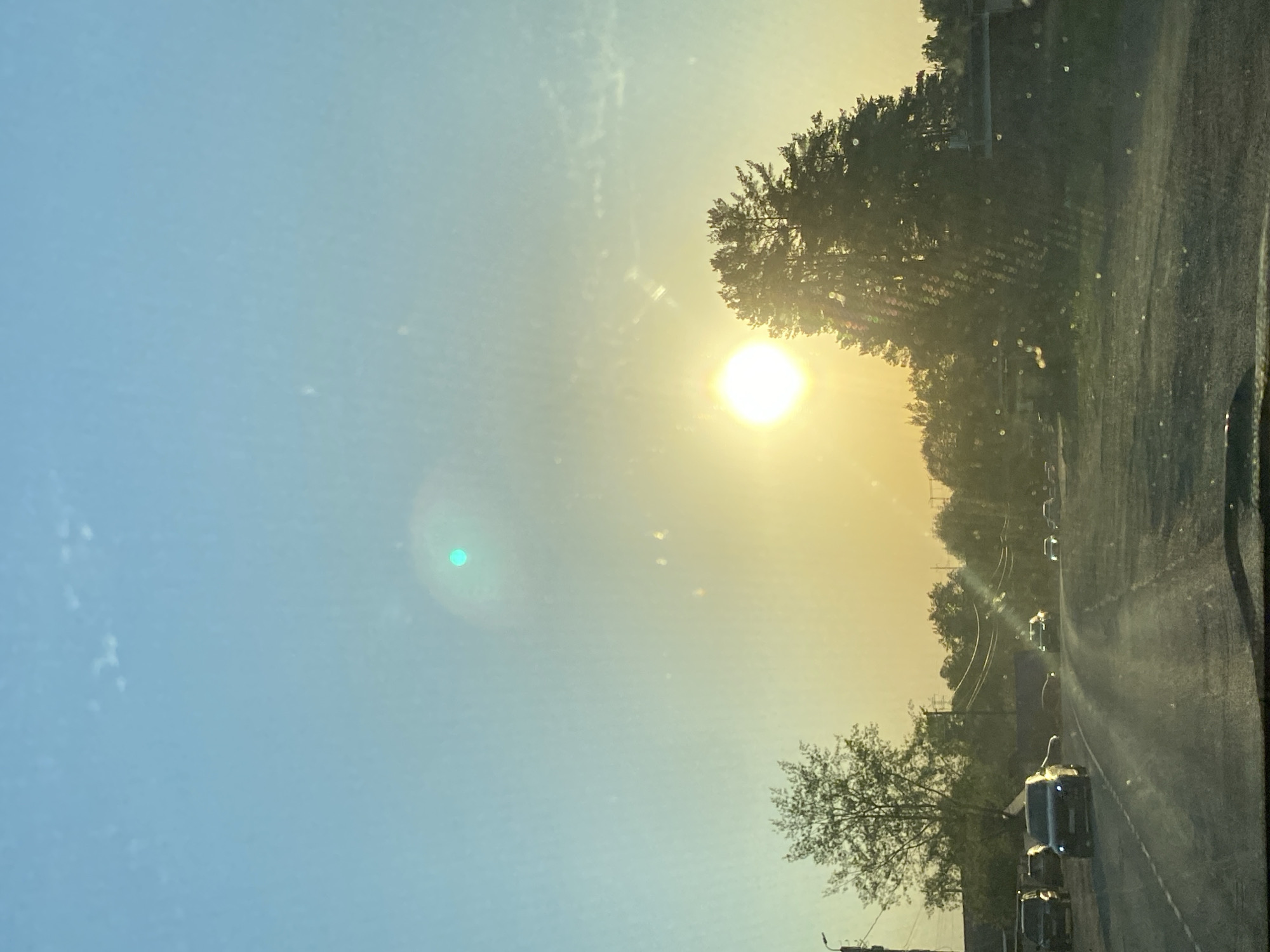 The image shows a view from inside a car, looking out onto a road with a bright sun in the sky. The sky transitions from blue to yellow due to the sunlight. There are trees and buildings visible along the roadside. Cars are driving on the road. The photo appears to be taken while driving, potentially in the city of Chaikovsky, Russia, if that information is accurate. The image lacks people and has a distinct focus on the environment and the road.