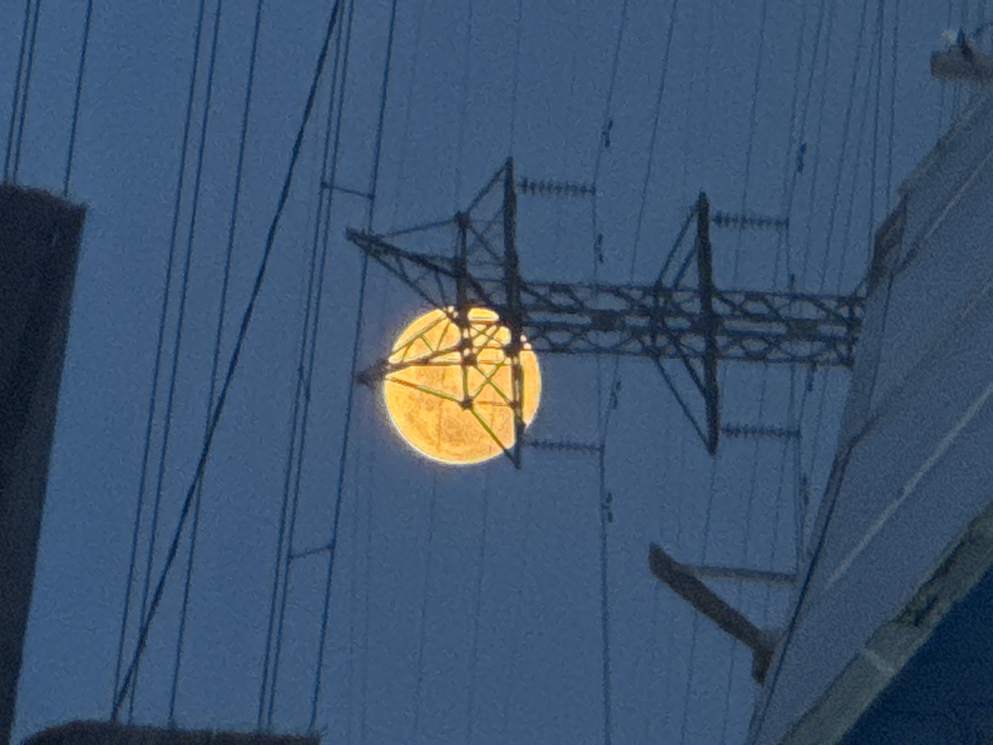 A low-angle, medium shot depicts a full, yellowish moon obscured by a power line structure against a dusky blue sky. The foreground contains partially visible buildings on either side, while the background is dominated by power lines that extend across the frame. The image was likely taken in an area with industrial or residential infrastructure, possibly within the city of Chaikovsky, Russia.