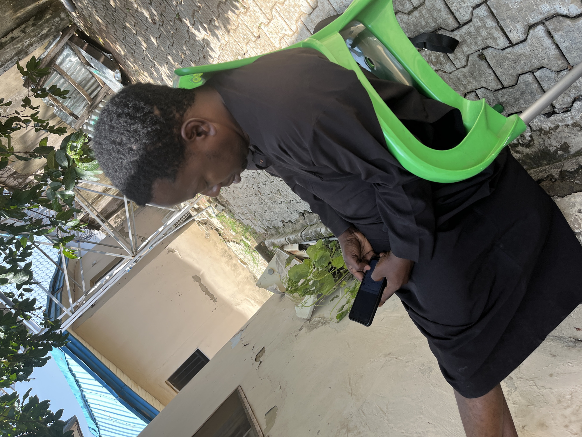 A person sits on a bright green plastic chair on a paved outdoor area. The individual, facing to the right and looking down at a smartphone, is dressed in dark clothing. The smartphone is held in both hands. The environment includes a building with a pale wall and a blue roof, and lush green foliage. The ground is covered with interlocking concrete pavers. The image appears to be taken in an outdoor setting within Galadimawa Village, Nigeria.