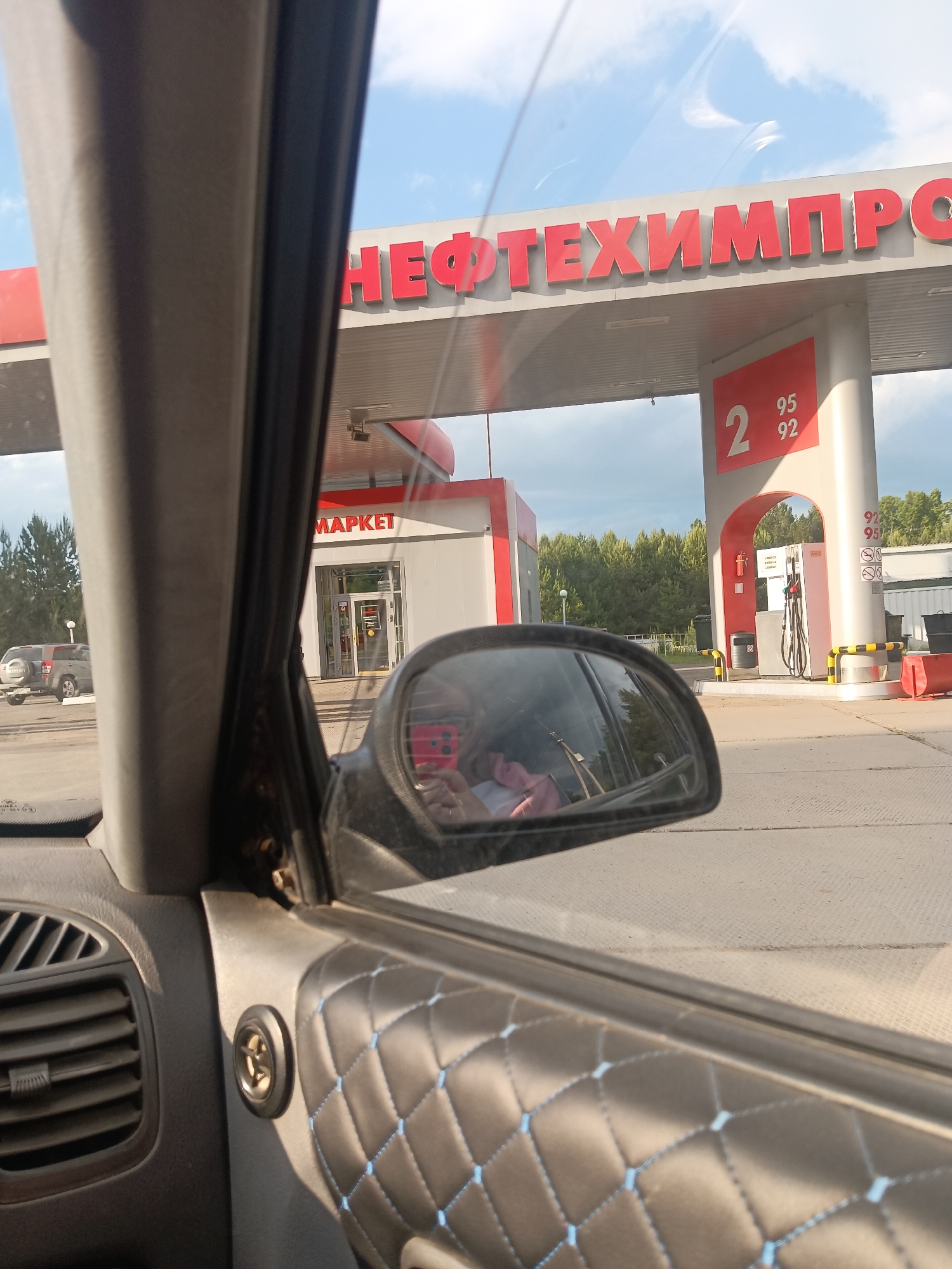 The image shows a view from inside a car at a "НЕФТЕХИМПРОМ" gas station. The location is likely in or near the city of Chaikovsky, Russia, based on the Cyrillic text on the gas station sign. The view through the driver's side window includes the gas station canopy, a market, and gas pumps. In the side mirror of the car, a person is taking a picture with a red phone. A Suzuki SUV is parked in the background near the market. The interior of the car is visible, featuring gray plastic and a quilted door panel with light blue stitching.