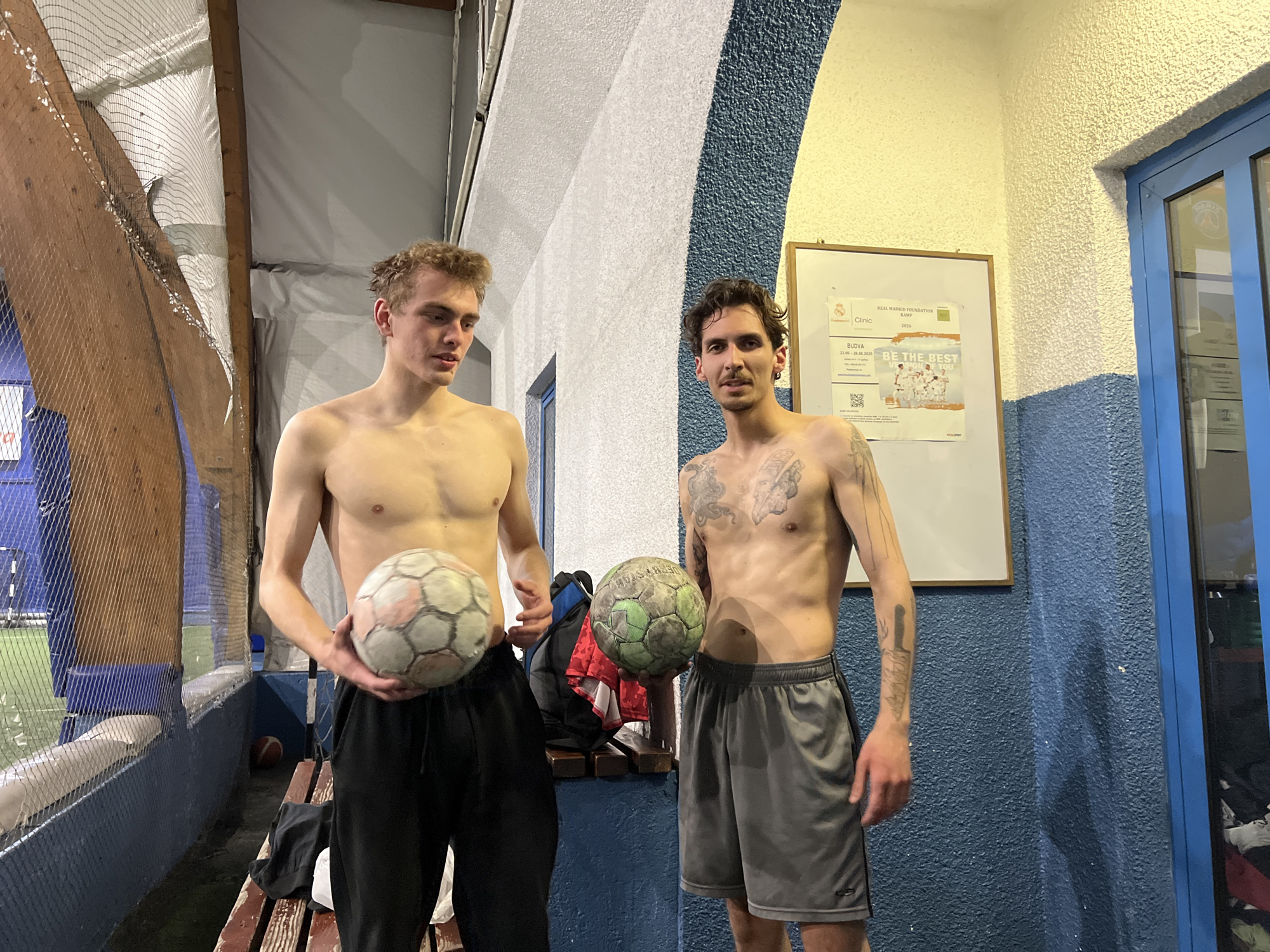 Two shirtless men stand indoors, each holding a soccer ball. The man on the left has blonde hair and wears black athletic pants. The man on the right has dark hair, visible tattoos on his torso and arms, and wears grey athletic shorts. Behind them is a wall, part of which is white with blue accents, and a bulletin board with a flyer for a "Real Madrid Foundation Kamp" Clinic in Budva, Montenegro, dated 22.06-28.06.2026. A door with a blue frame is visible to the right of the man with tattoos. To the left of the man with blonde hair is a netted area, suggesting an indoor soccer field or similar sports facility. A wooden bench with clothing on it is behind the men.
