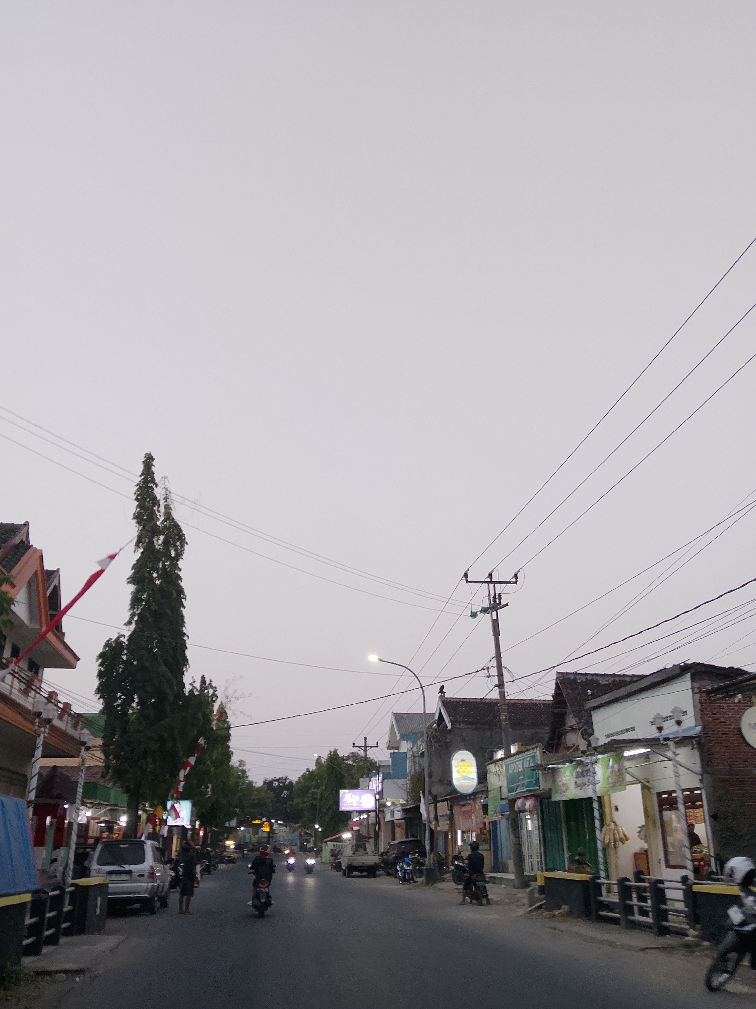 The image depicts a street scene in Ngelo, Indonesia, likely in the late afternoon or early evening, given the dim lighting and the overcast sky. The street is paved and appears to be a local thoroughfare with buildings lining both sides.
On the left side of the street, there are several buildings, including a structure with orange and white accents on its roof. A tall, slender tree stands prominently in front of these buildings, with a red and white flag (likely the Indonesian flag) attached to a pole near its base. A silver SUV is parked on the side of the road.
The right side of the street features a row of shops and businesses. Some of the signs are illuminated, suggesting they are open. One sign appears to say "APOTEK KITA," indicating a pharmacy. There are awnings and various architectural styles among the buildings, suggesting a mix of older and newer constructions.
Several people and vehicles are present on the street. Motorcycles are a dominant mode of transport, with several individuals riding them. Two people are standing on the sidewalk near the parked SUV. A person is visible inside one of the buildings on the right. Power lines and utility poles are visible throughout the scene, crisscrossing the sky. The overall impression is of a moderately busy, local street in a residential or commercial area.