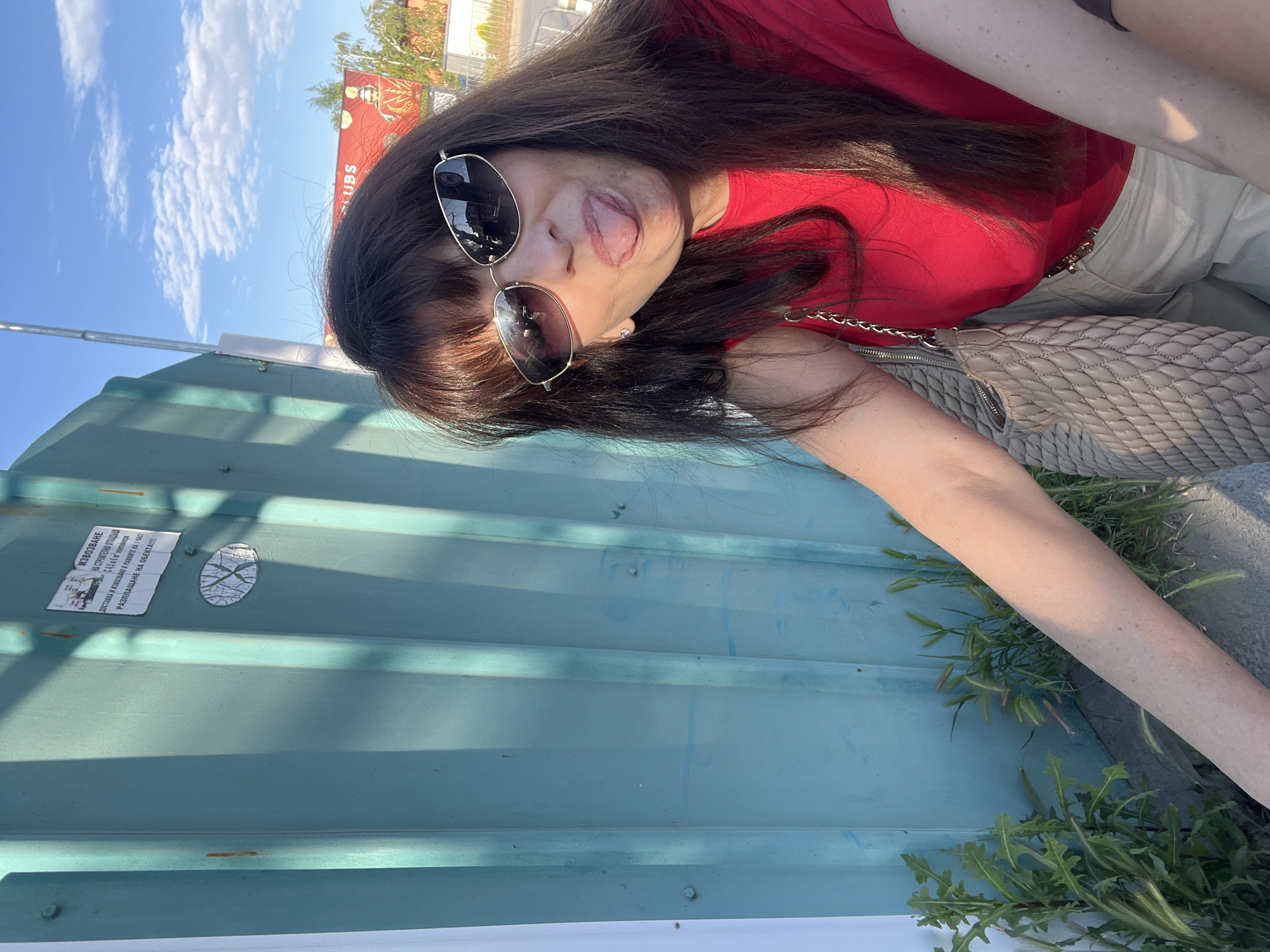 A woman with sunglasses, a red shirt, and a light-colored bag is sticking her tongue out at the camera. She is standing in front of a blue metal structure with Russian writing on a sticker. The sky is visible in the background. A portion of an advertisement for "UBS" is visible above her head. Based on the text in the ad and the Russian writing, the location is likely in Plovdiv, Bulgaria. Greenery is growing near the bottom of the metal structure.