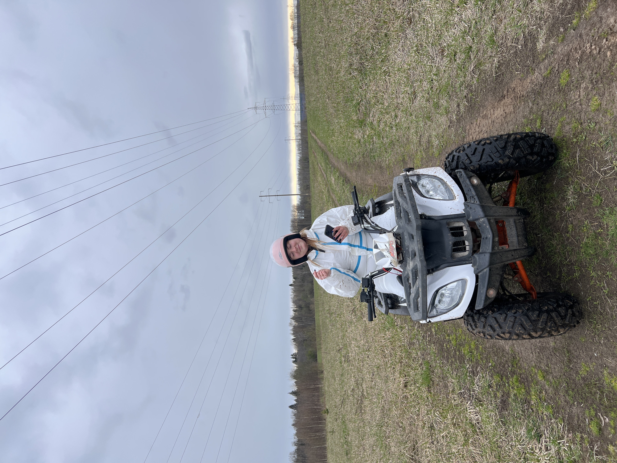 A woman in a white jumpsuit and pink helmet sits on a white ATV in a grassy field, holding a smartphone. Overhead power lines run across the sky to a transmission tower in the distance. Trees line the horizon. The location is near Razdolye, Russia.