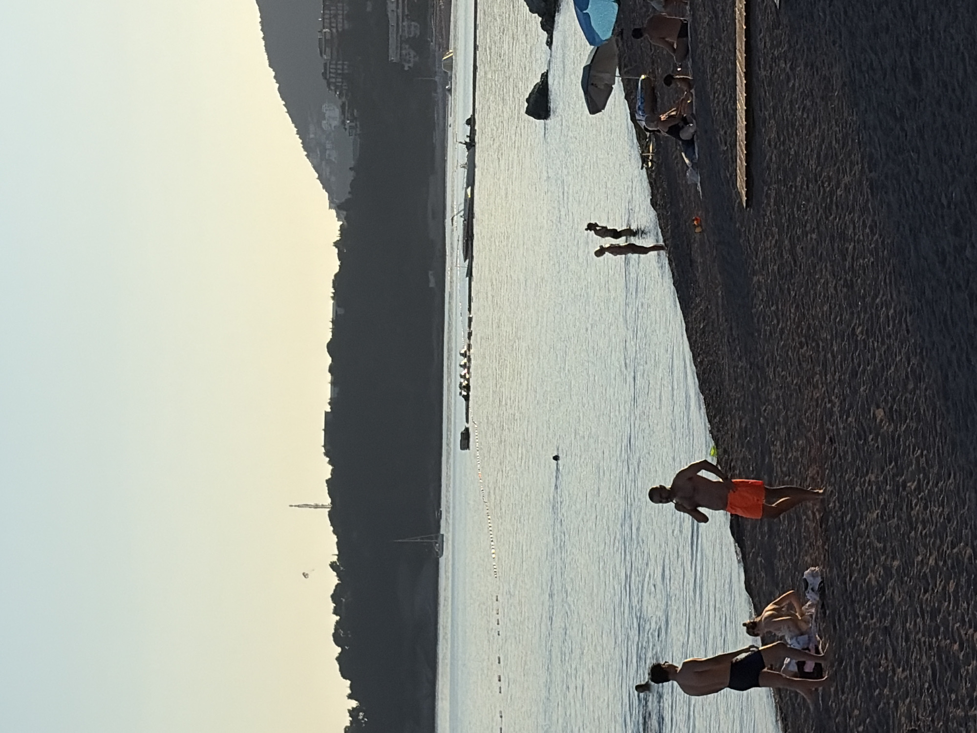 The image depicts a beach scene in Rafailovići, Montenegro, along the Adriatic coast. The scene captures people enjoying various activities. Along the beach, the sand slopes down to the water's edge. Several individuals are seen on the beach; some are in the water, swimming or wading, while others are on the shore, possibly resting or sunbathing. The individuals closer to the shoreline are clearly visible, showcasing their attire. Behind the beach, vegetation densely covers the mountainous terrain. Further into the sea, a string of buoys marks off a swimming area. In the distance, buildings and possibly more of the city are visible, adding depth to the view. The sky is light, suggesting either early morning or late afternoon.