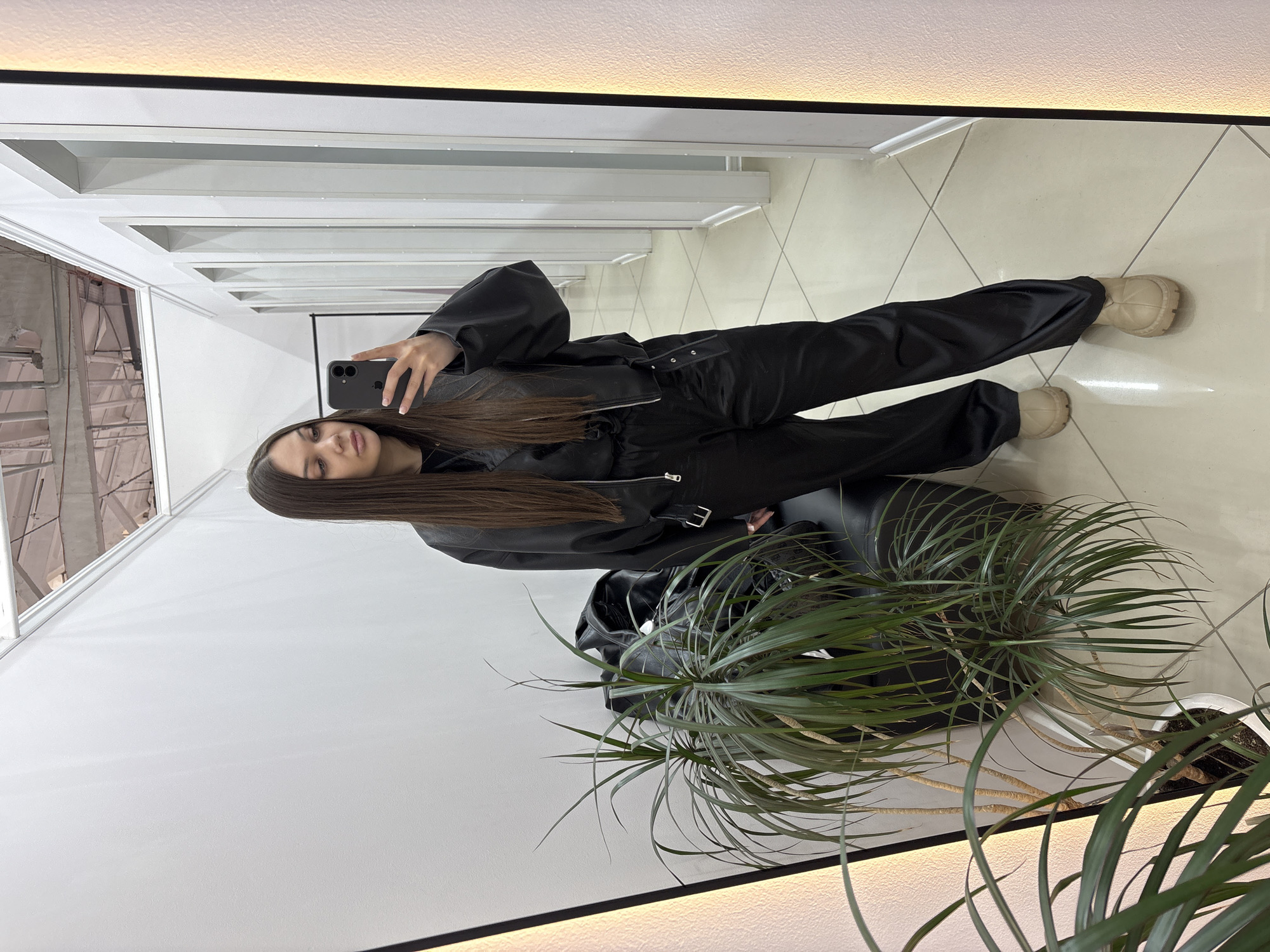 A woman stands in front of a full-length mirror in what appears to be an indoor space, possibly a business establishment in Chaikovsky, Russia. She is holding a smartphone, presumably taking a photograph of herself. The woman is wearing a black jumpsuit and beige platform shoes. The room features white walls, white horizontal architectural elements near the ceiling, and a tiled floor. There is a potted plant with long, thin leaves in the foreground. A black chair is visible behind the plant.
