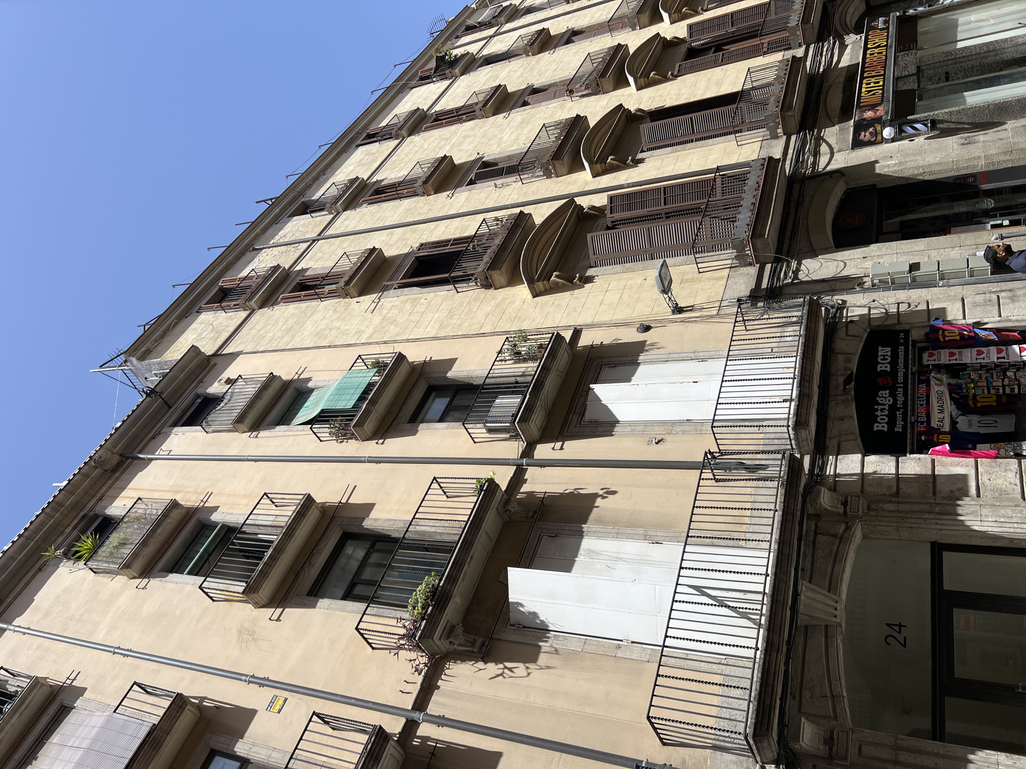 The image shows the facade of a tan-colored, multi-story building under a clear blue sky. The building features several balconies with black metal railings, some with potted plants and greenery. Many windows are visible, some with closed brown shutters and others with blinds. The lower part of the building has shops with signage. One sign reads "Botiga BCN Esport, regals i complements" with FC Barcelona merchandise visible. Another sign says "MISTER BARBER SHOP". A street lamp is attached to the building. A person is partially visible in the lower right corner. The architecture and storefronts suggest an urban setting, consistent with a street in Barcelona, Spain.
