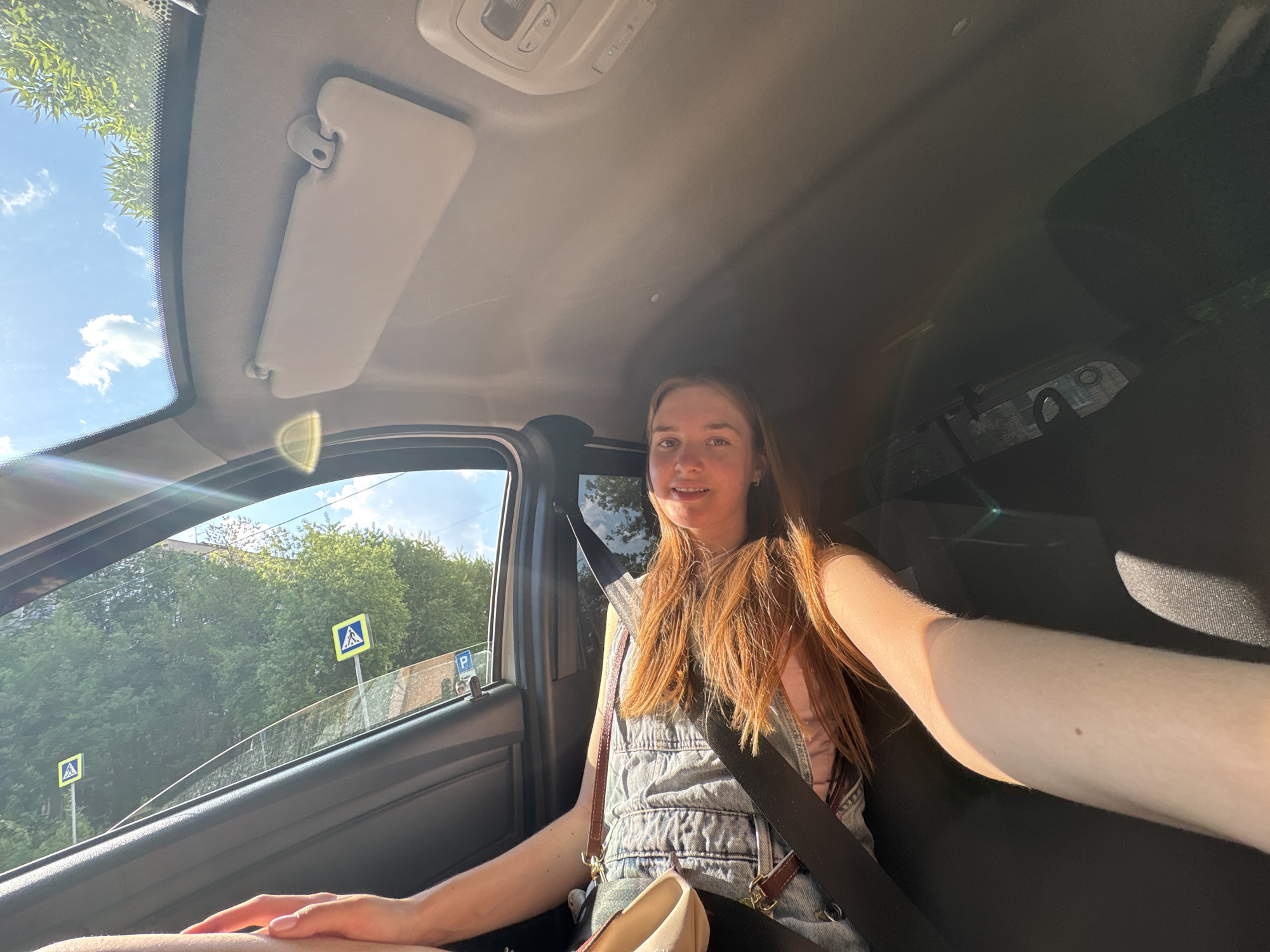 A fair-skinned young woman with long, reddish-brown hair is seated in the driver's seat of a car, wearing a light-colored denim overall dress and a seatbelt. She appears to be taking a selfie with her right arm extended toward the camera. Through the car window, the streetscape suggests a location in Perm, Russia, with trees and a pedestrian crossing sign visible. The interior of the car is visible, including the sun visor, dashboard, and seating.
