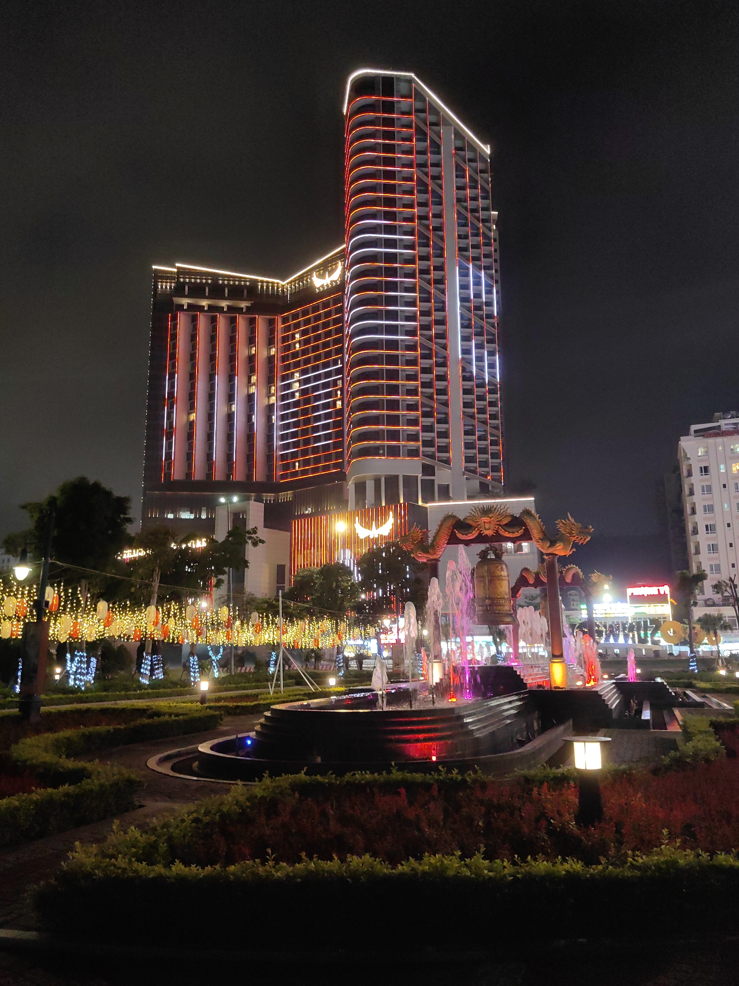 At night in Bai Chay Ward, Vietnam, a multi-tiered fountain with colorful lights illuminates a landscaped garden. A golden bell hangs within a dragon-adorned structure behind the fountain. In the background rises a tall hotel, its facade lined with red and white lights. A row of lanterns hangs across the foreground.