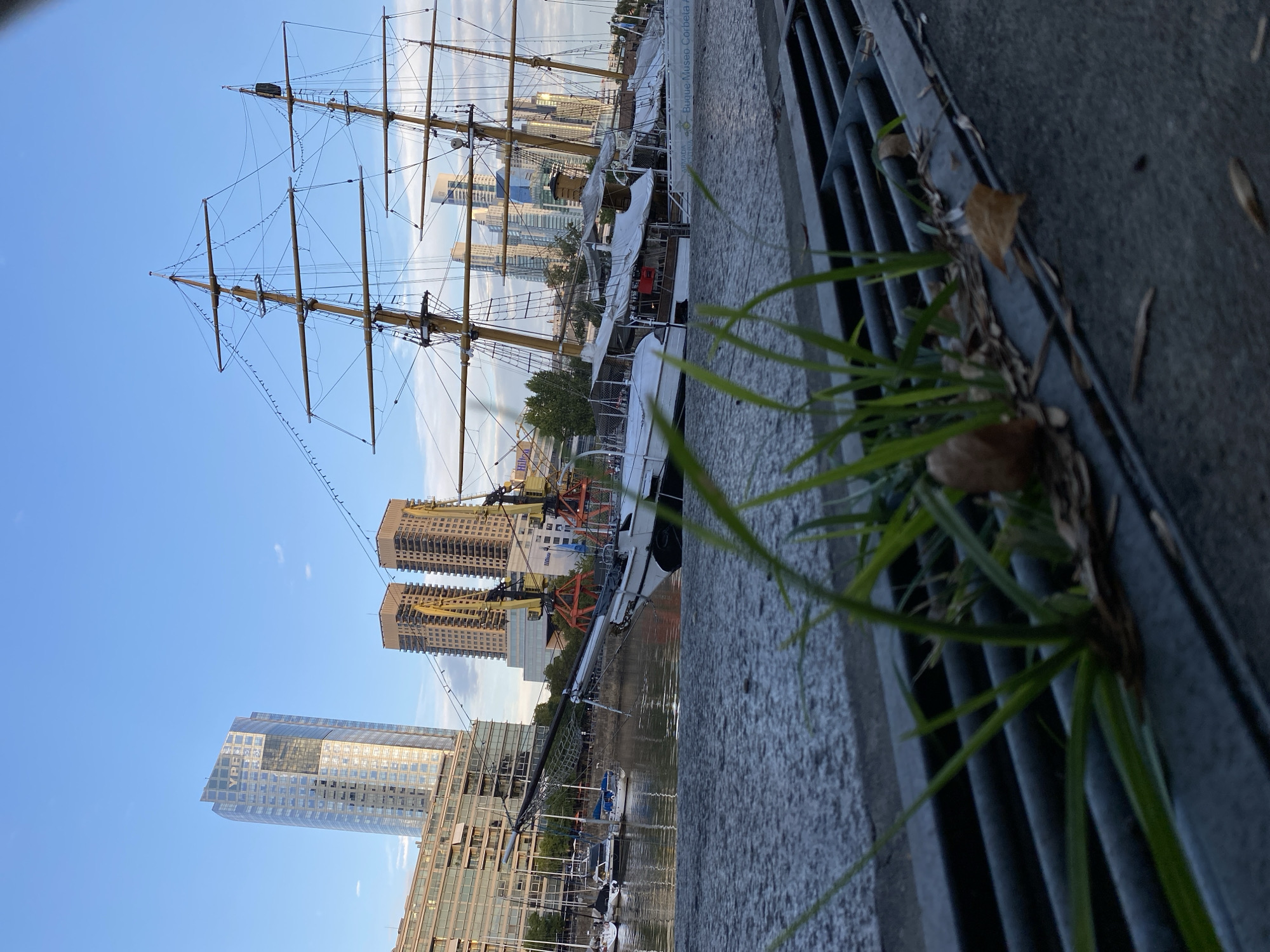 A slightly angled, wide shot taken near the dock of Puerto Madero, Buenos Aires, Argentina. Tall buildings line the waterfront. A large, tall ship with masts occupies the center. Along the bottom right, there is a cement barrier and a metal grate. Some green grass and dried leaves grow near the grate. There are other smaller boats docked along the water.