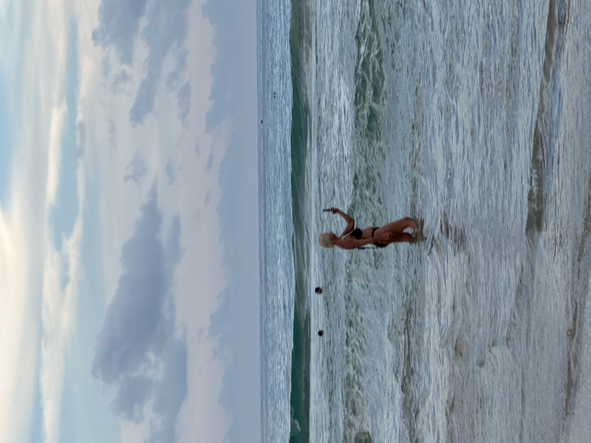 At Karon Beach in Karon, Thailand, a woman with blonde hair stands in the ocean, wearing a black bikini, and takes a selfie with her phone. The water is choppy with small waves breaking around her. In the background, two other individuals are visible further out in the sea, with their heads above the water. The sky is overcast with scattered clouds.